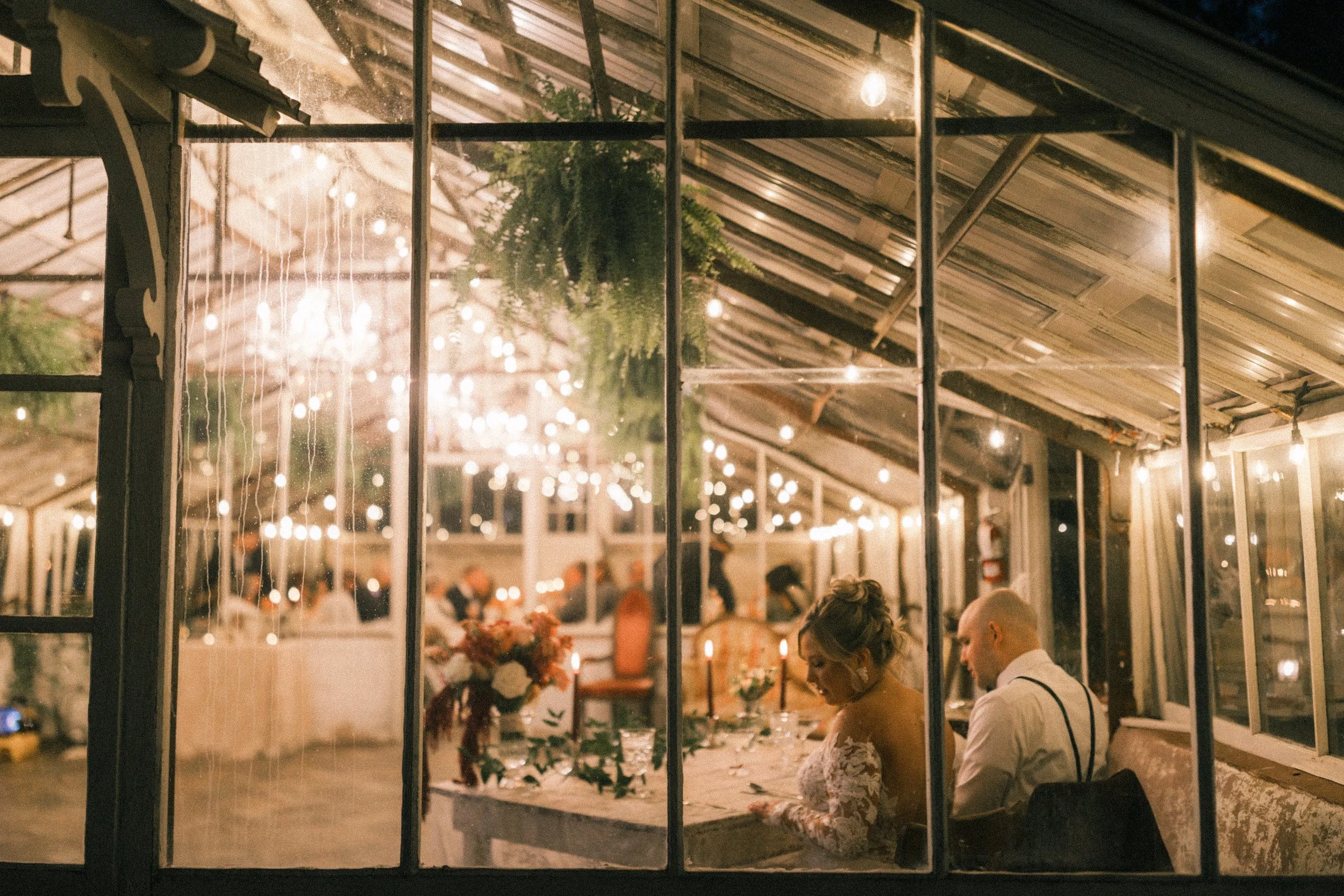 A cozy indoor event space viewed through a large window with a black metal grid. Inside, string lights, greenery, and festive decorations create a warm, inviting atmosphere. Guests are seated at tables, and a woman and man are engaged in conversation.