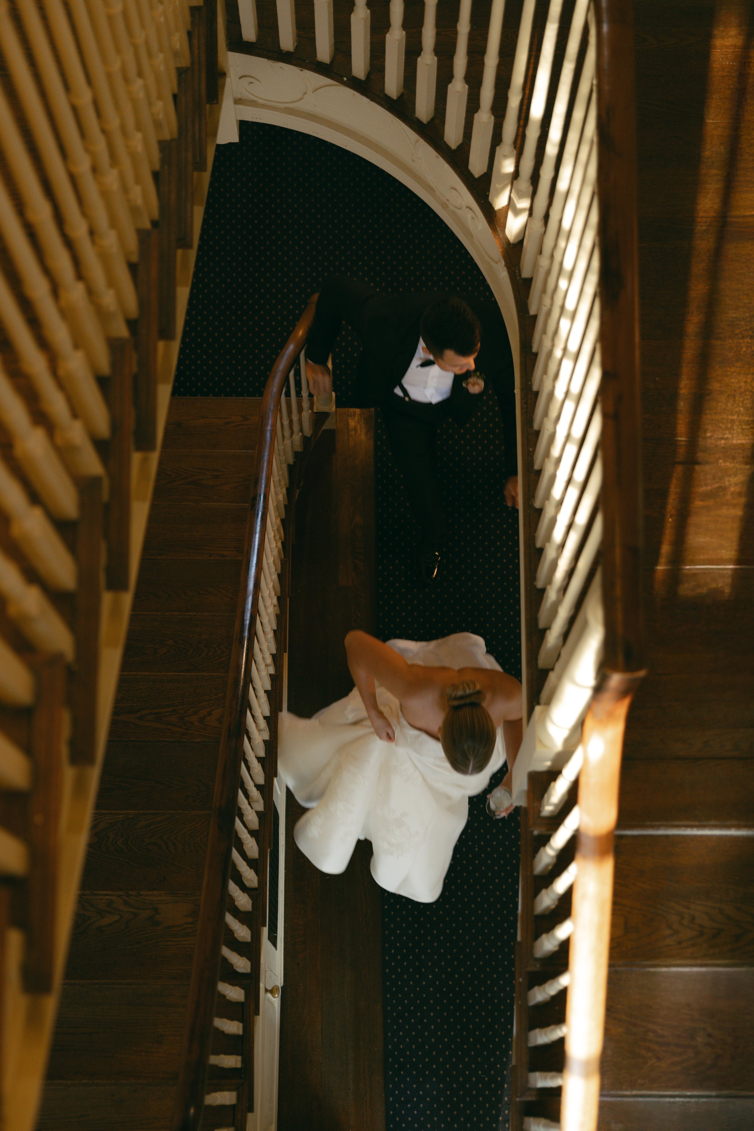This is a view from above of a bride in a white wedding dress lying on a bed, with a groom in a black tuxedo leaning over her, taken from a staircase.