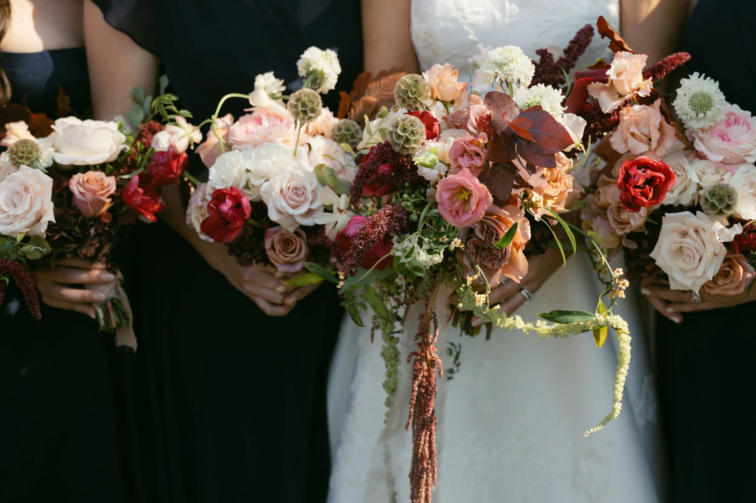 Close-up of a bridal bouquet with various pink, white, and red flowers held by people dressed in black and white attire.