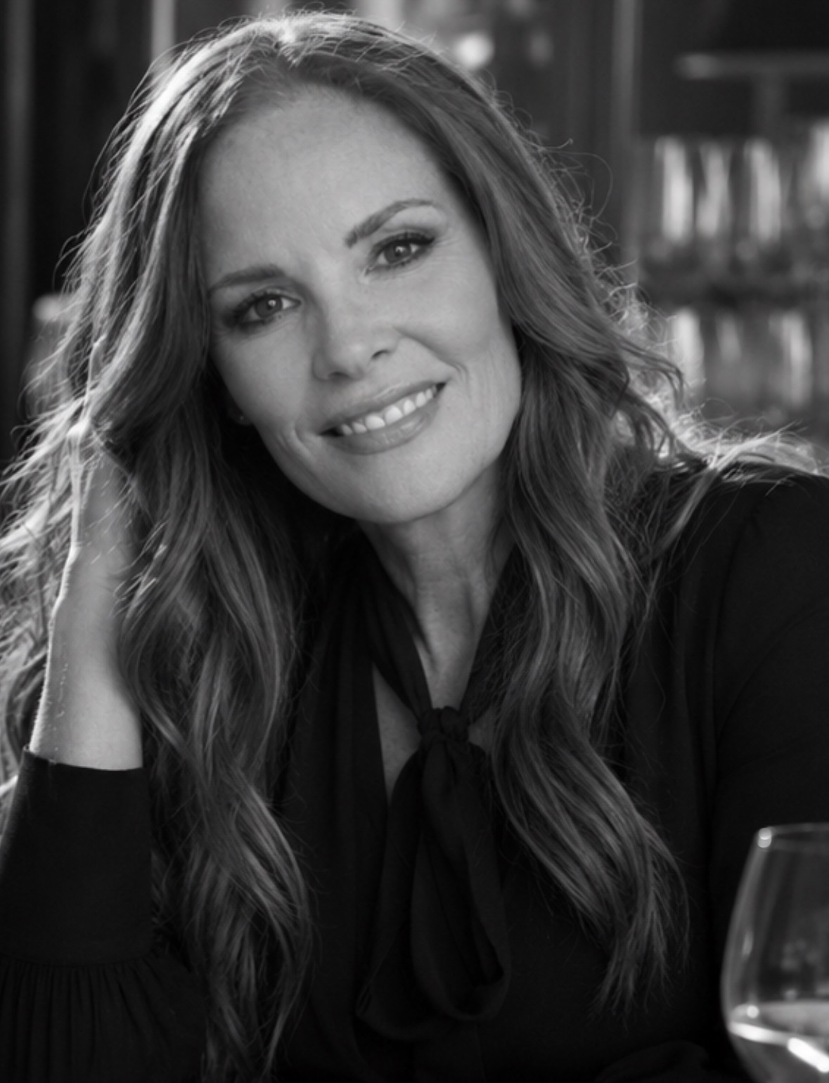 Black and white photograph of a woman with wavy hair smiling at the camera, seated at a table with a wine glass in front of her.