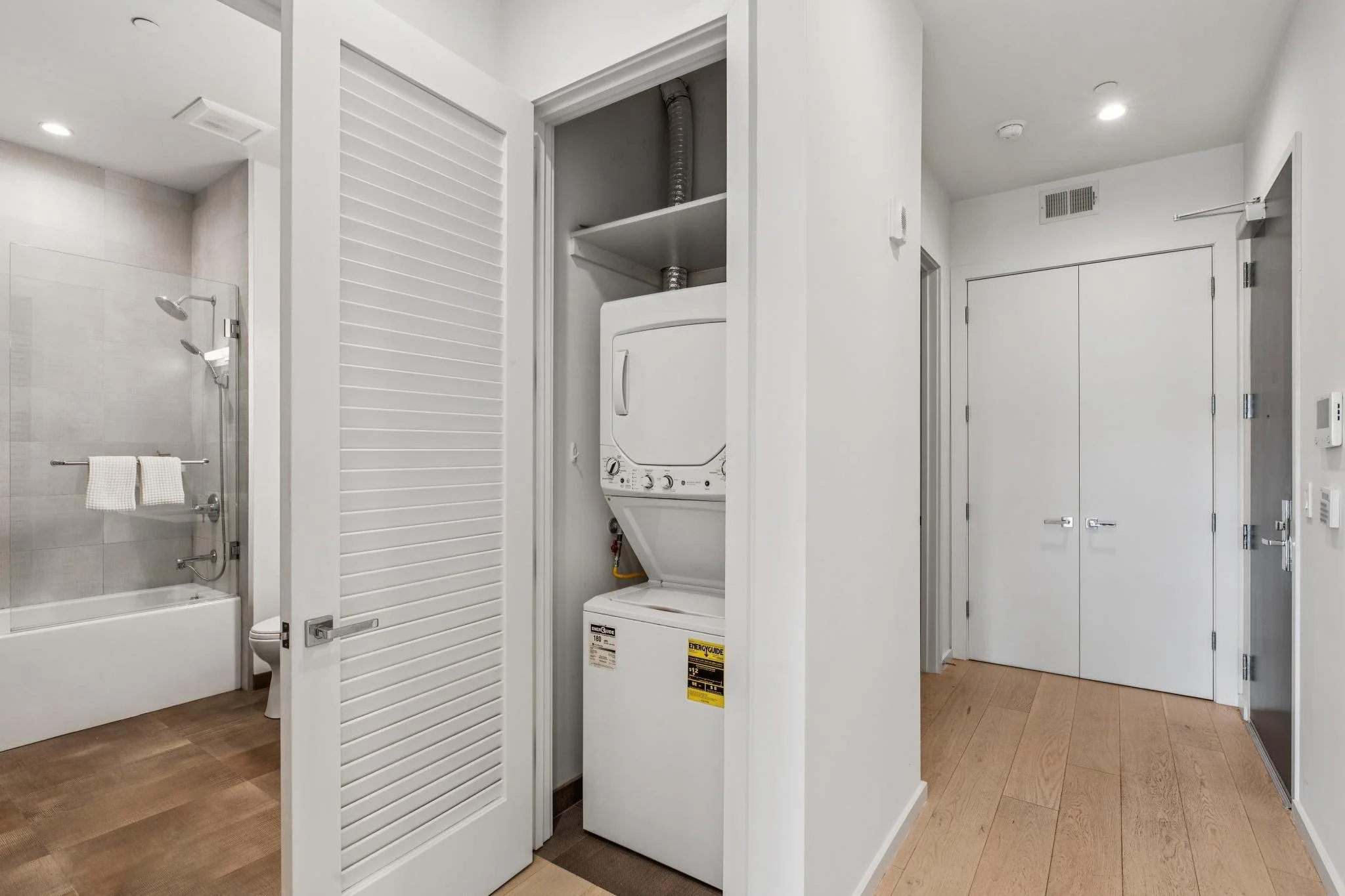 A laundry area with a stacked washer and dryer behind a partially open louvered white door, with a bathroom visible on the left and a hallway with double doors on the right.