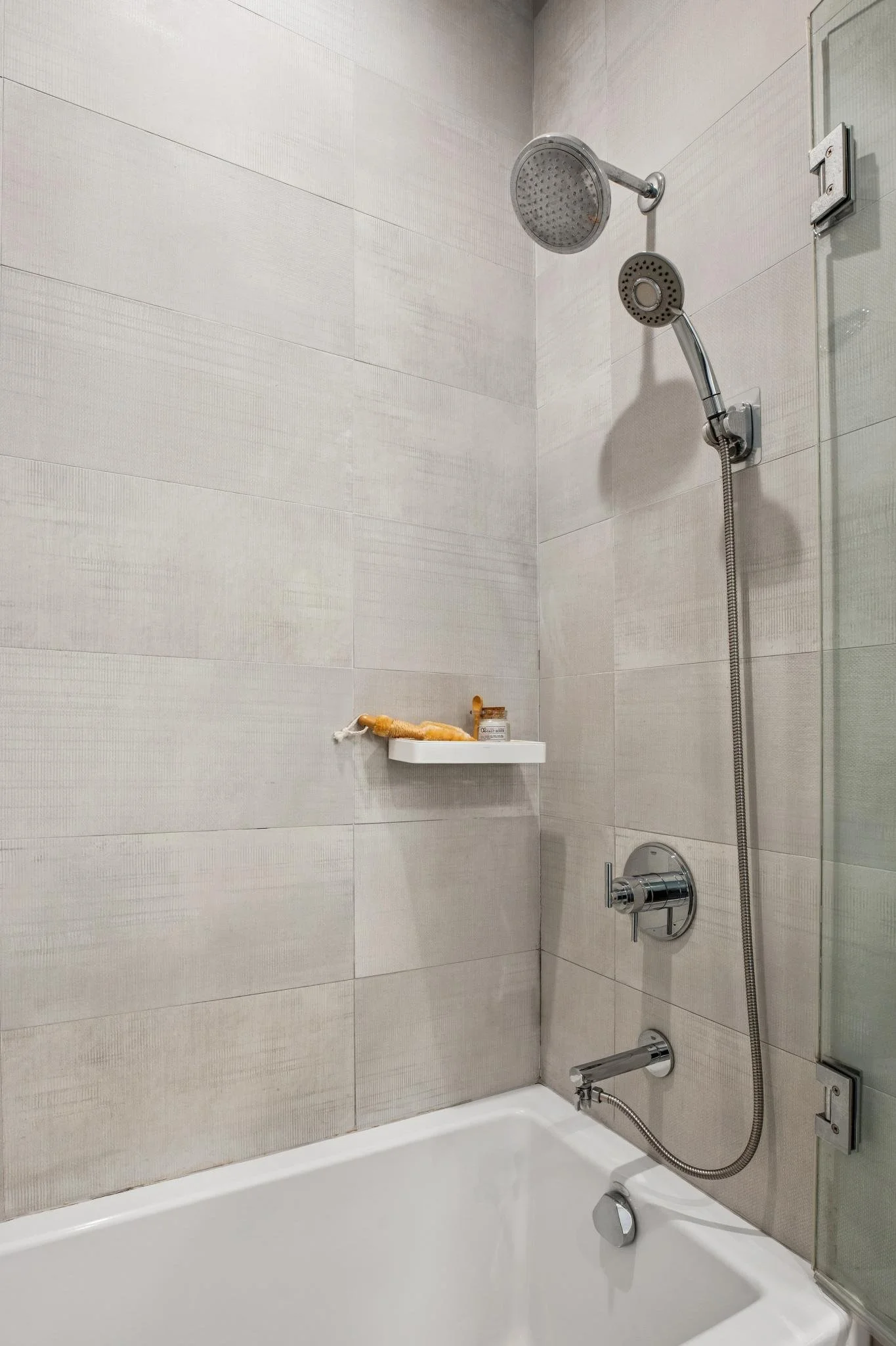 Shower with chrome showerhead and handheld sprayer on tiled wall, small white shelf with soap and shampoo, bathtub with silver faucet.