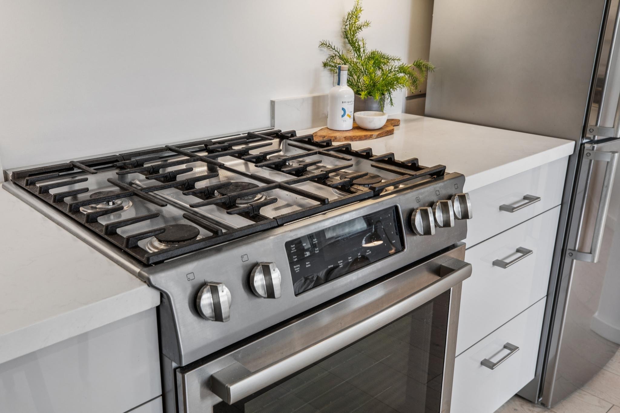A modern kitchen stove with a white countertop, a potted plant, a bottle, a bowl, and a cutting board on the counter.