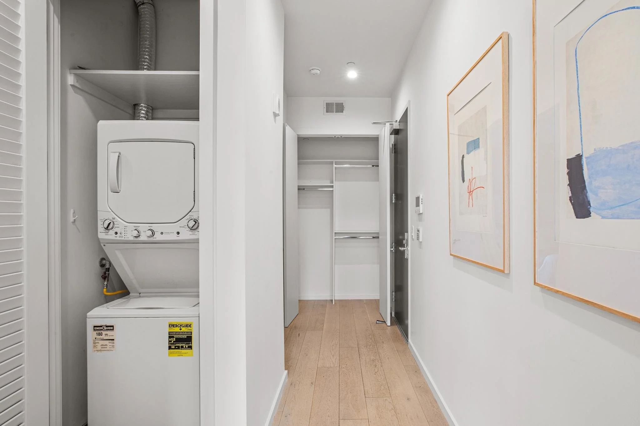A small laundry closet with a stacked white washer and dryer, and an open closet space with shelves and a hanging rod, all in a modern apartment hallway with light wood flooring and white walls with colorful abstract artwork.