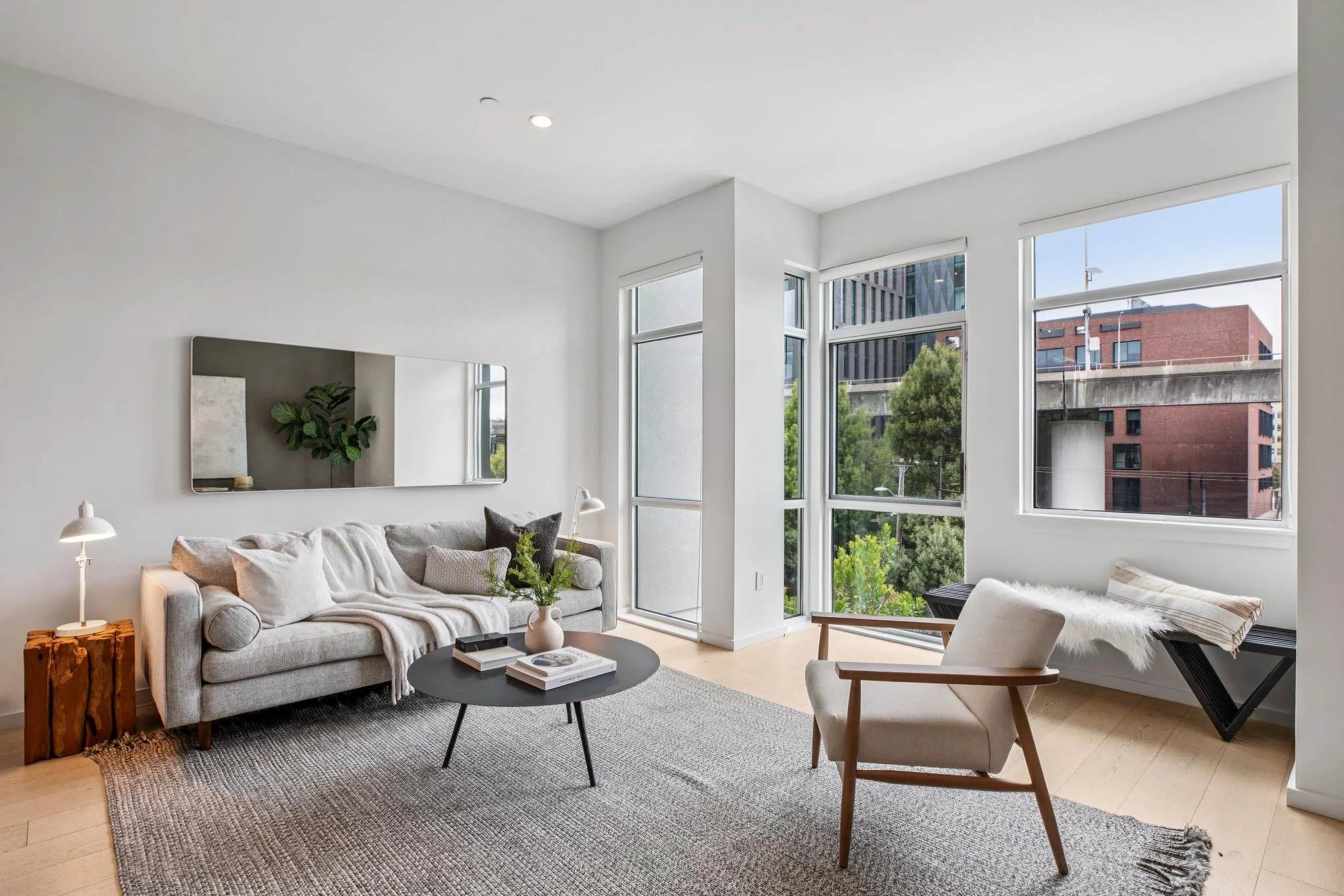 A living room with large windows, a gray sofa with white pillows, a round black coffee table with magazines, a wooden side table with a white lamp, a white armchair with wooden arms, and a large mirror reflecting a plant, all inside a modern apartment with wood flooring and light walls.
