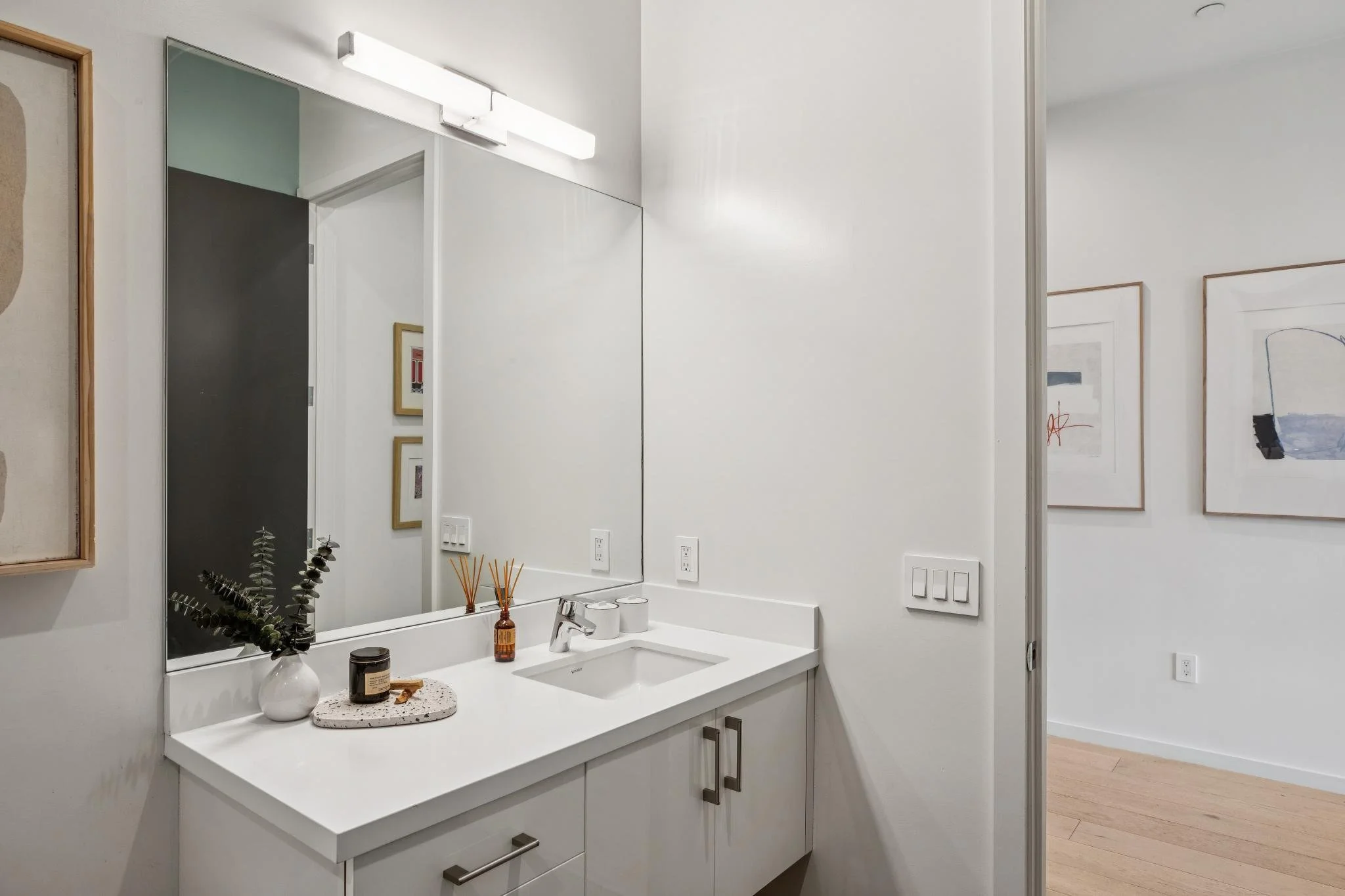 A modern bathroom with a white vanity, rectangular mirror, and decorative items including a vase with eucalyptus, a candle, and a small tray. The walls are white, with framed artwork visible in the reflection.