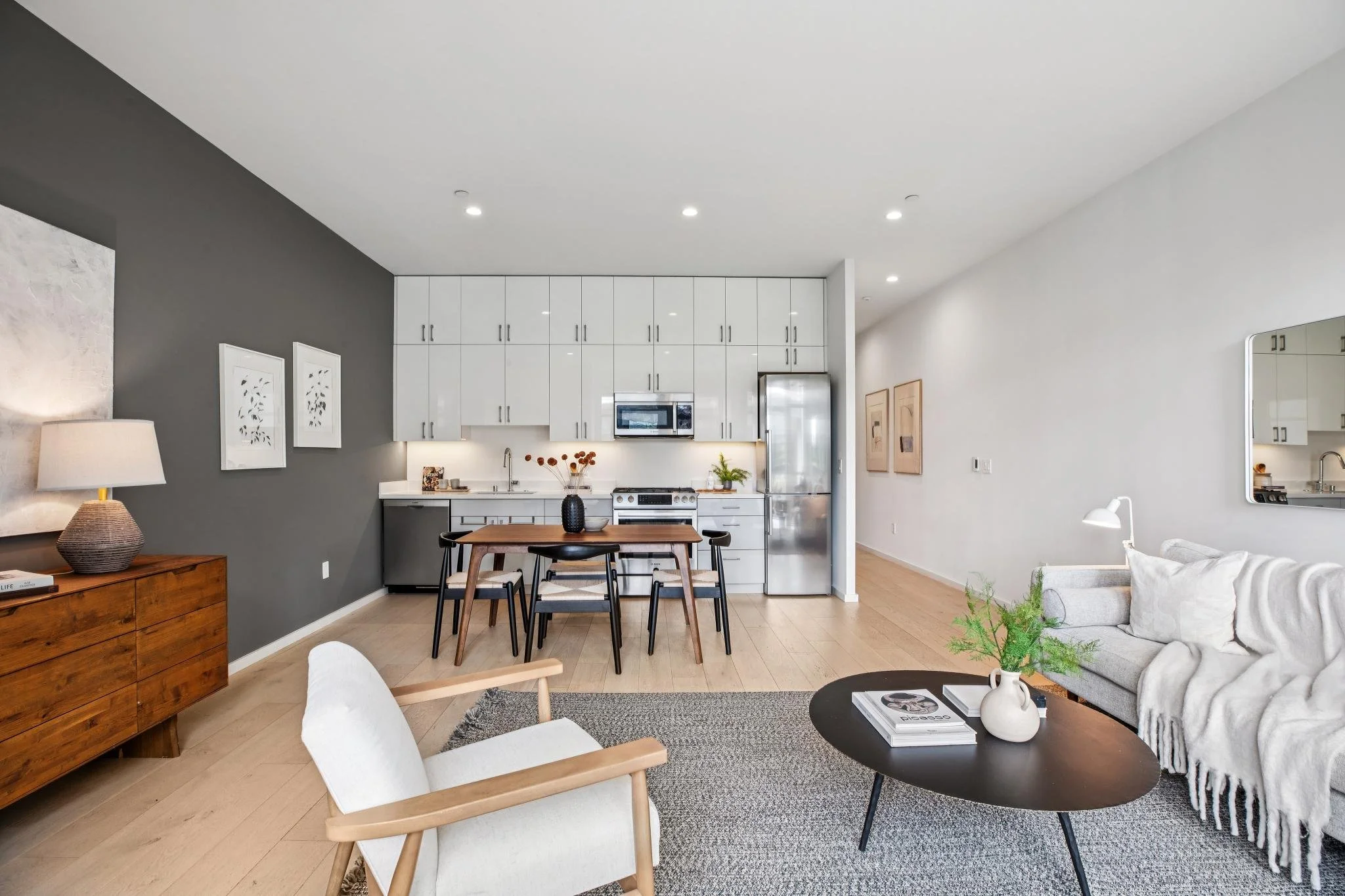 Open-concept living room and kitchen in a modern apartment with white and gray walls, light wood flooring, white cabinetry, stainless steel appliances, a black round coffee table with a white vase and books, a cozy white sofa with pillows, and a wooden armchair with a beige cushion.