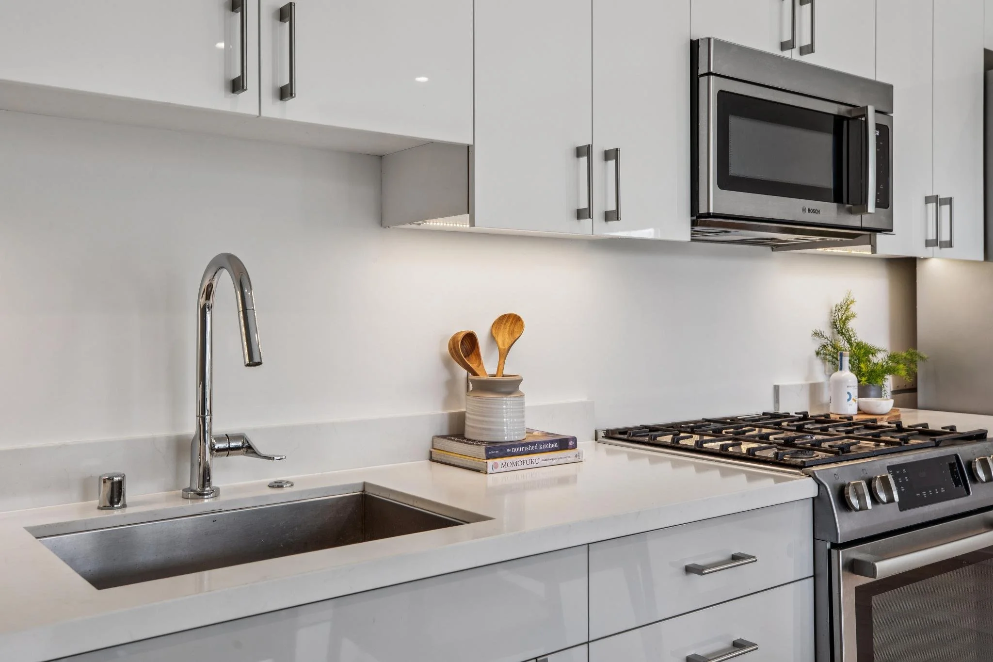 Modern kitchen with white cabinets, stainless steel microwave, and stove. Kitchen counter holds a stack of books, a decorative plant, and wooden spoons.