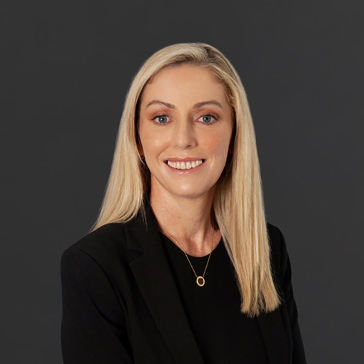 A woman with long blond hair smiling, wearing a black blazer and a gold necklace, posed against a dark gray background.