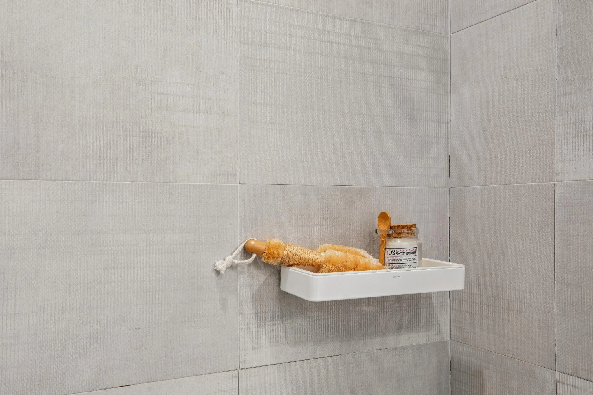 A corner of a bathroom with light gray textured tiles on the walls. There is a white wall-mounted sink with a wooden scrub brush resting on top, a small bottle of salt scrub, and a loofah hanging from the side.