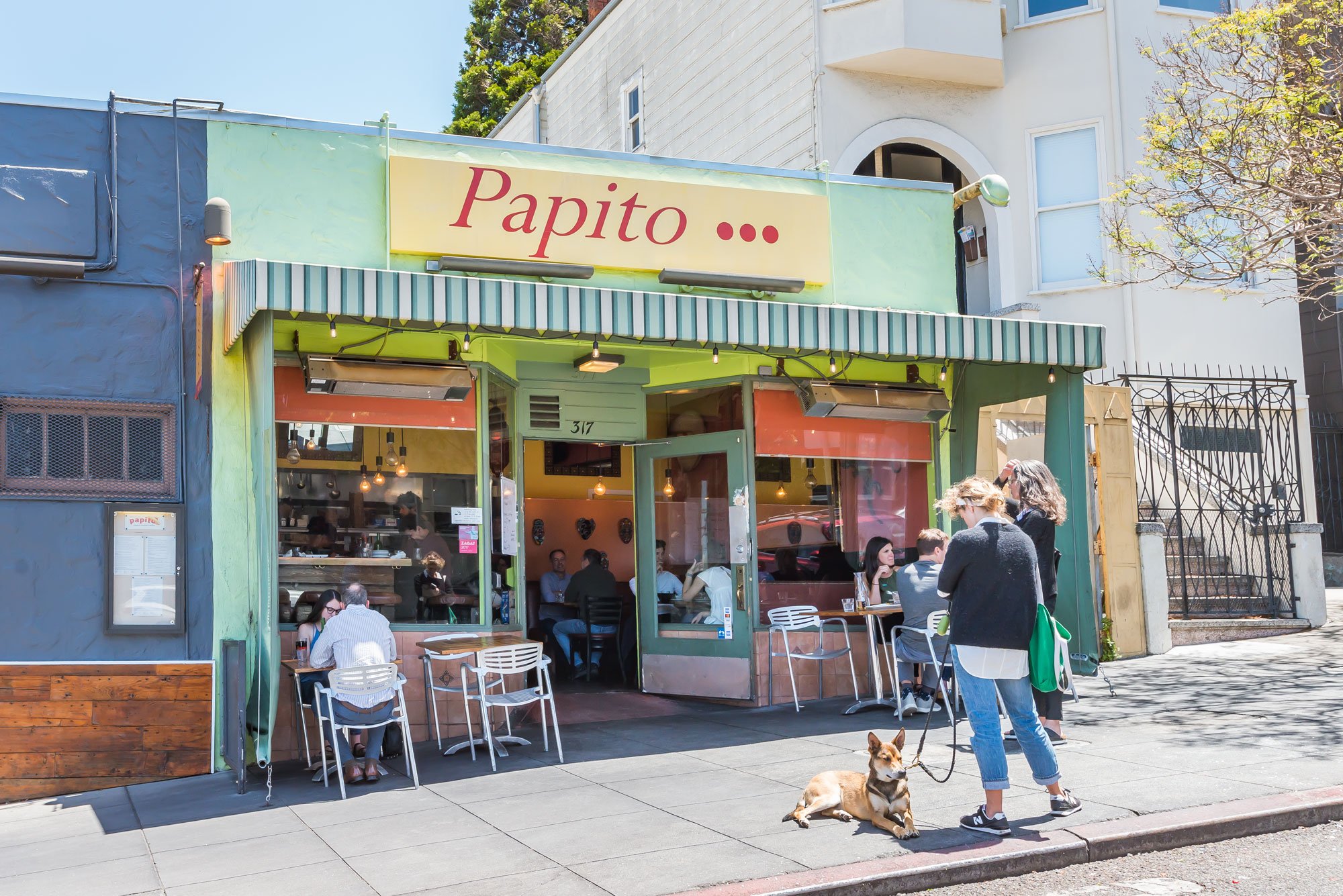 Exterior view of a restaurant called Papito with people dining inside and outside, and a woman walking her dog on the sidewalk during daytime.