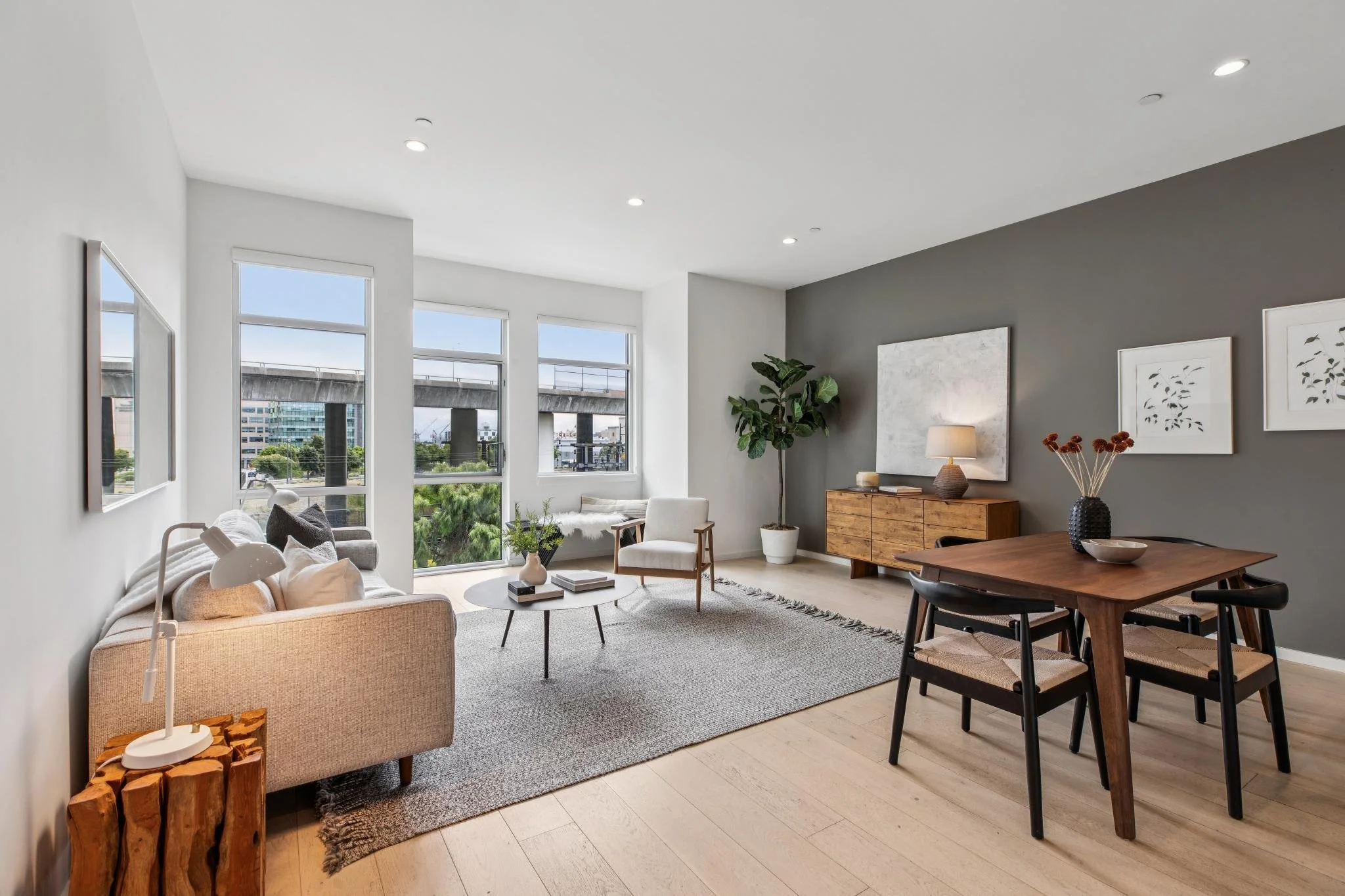Modern living room with large windows, white walls, a beige sofa, a wooden coffee table, a white armchair, plants, and wall art, with a view of a cityscape.