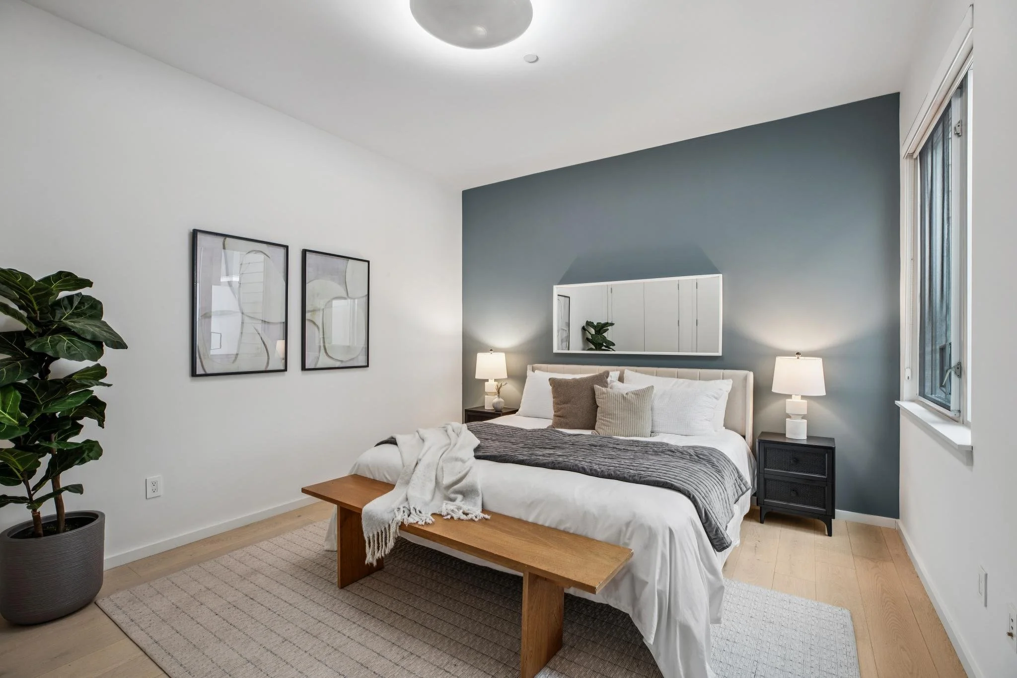 A modern bedroom with a white bed, gray blankets, and multiple pillows. There is a large potted plant on the left, two black nightstands with white lamps, and two framed abstract art pieces on the white wall. A wooden bench sits at the foot of the bed, and a window on the right fills the room with natural light.