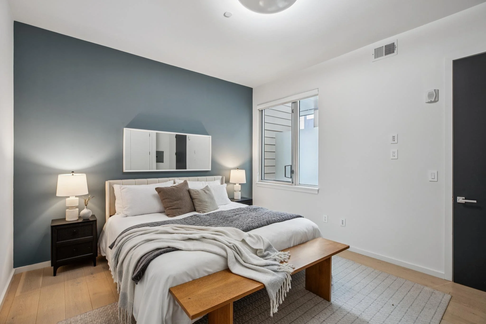 Minimalist bedroom with white and gray color theme, a bed with white linens and brown pillows, black nightstand with two white lamps, framed mirror on the gray accent wall, window allowing natural light, light wood flooring, and white walls.