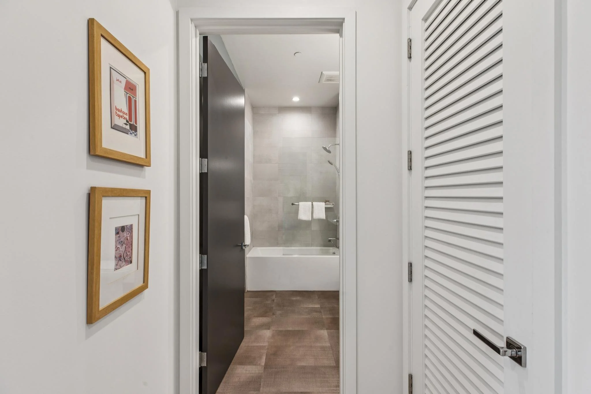 View of a bathroom seen through a doorway from an adjacent room with three framed pictures on the white wall to the left. The bathroom has a shower with beige tiles, a showerhead, and a tub, with a towel hanging on a rod below the shower.