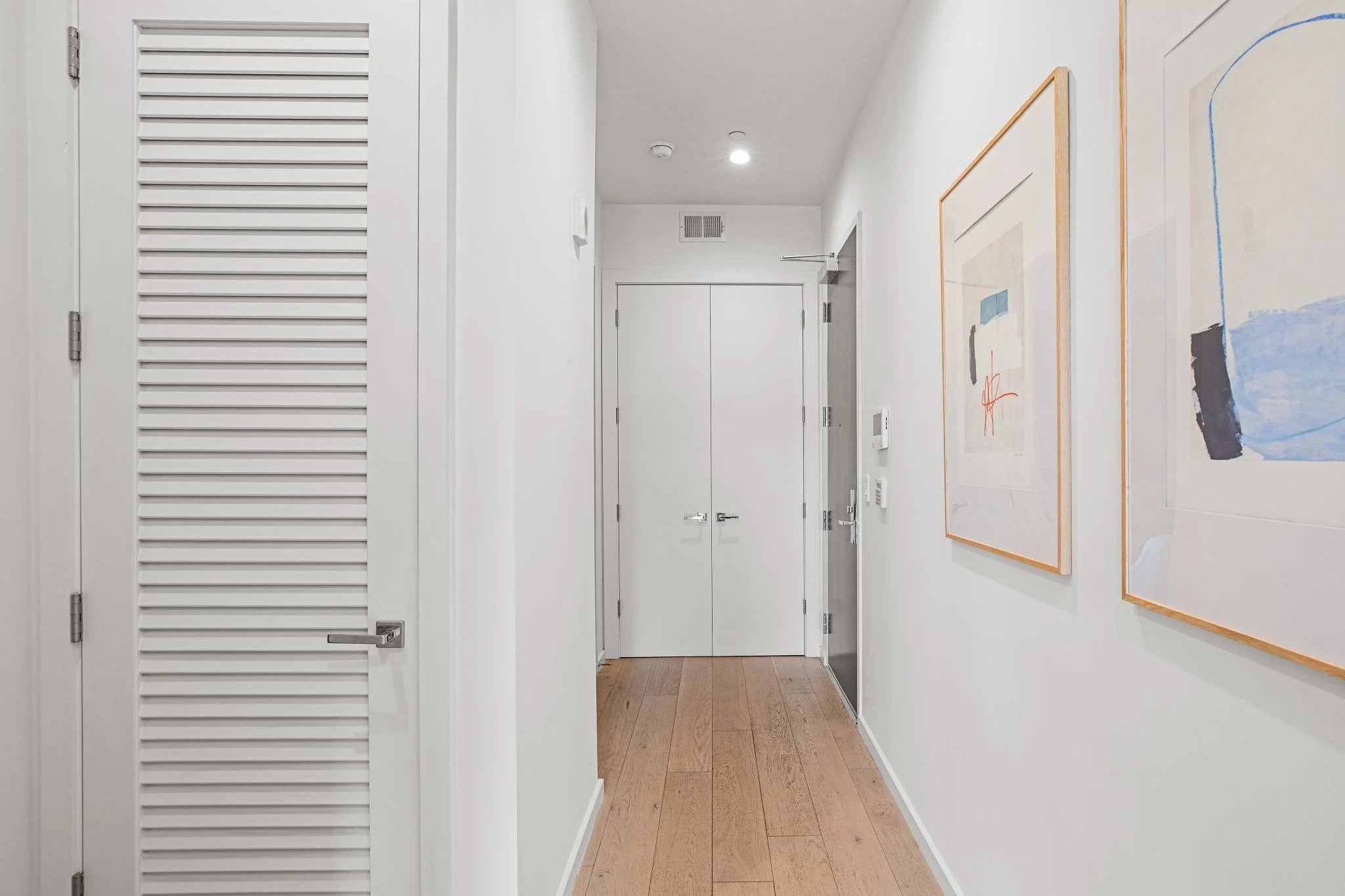 A hallway with white walls, wooden flooring, and two abstract paintings hanging on the right wall. There are closed double doors at the end of the hallway, and a smoke detector and some wall-mounted devices are visible on the walls.