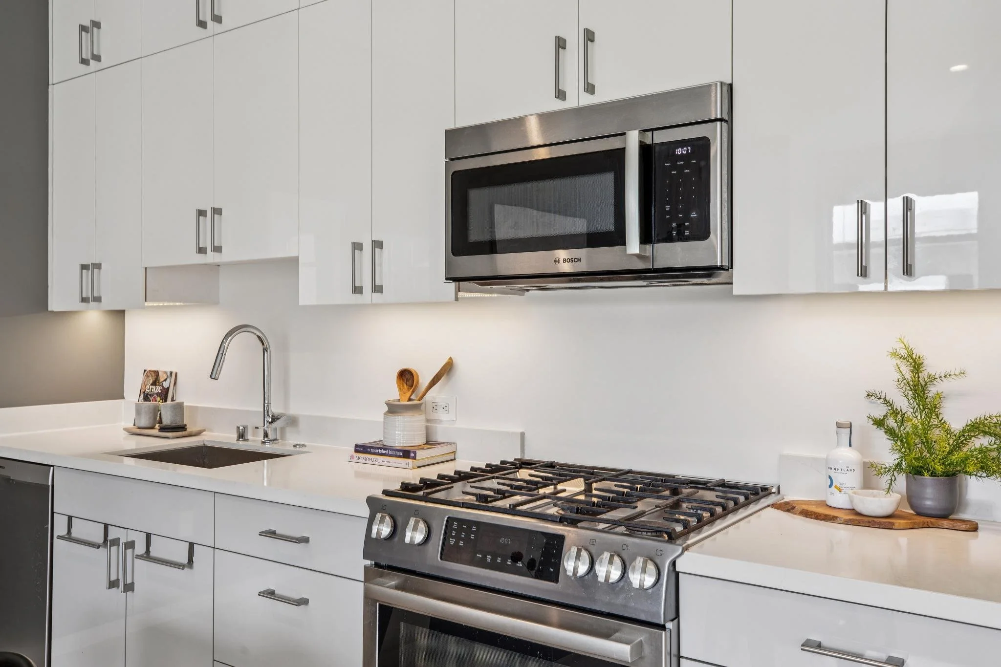 Modern kitchen with white cabinets, stainless steel Bosch microwave above a gas stove, white countertop, sink, and decorative items including books, a potted plant, and utensils.