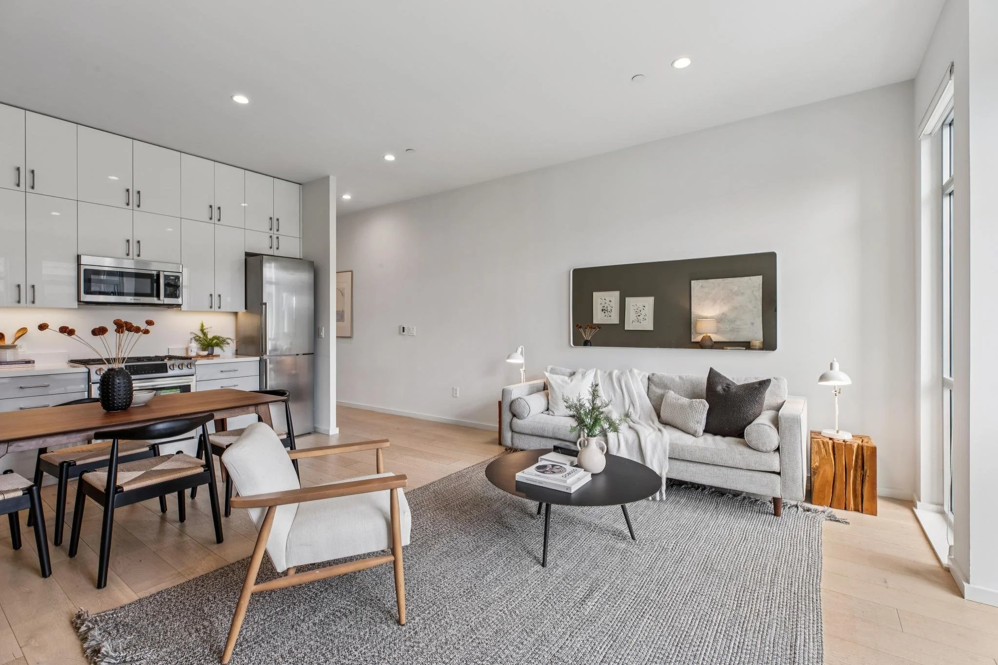 Modern living room with white walls, beige sofa, black coffee table, and dining area with wooden table and black chairs. Kitchen in background with white cabinets and stainless steel appliances.