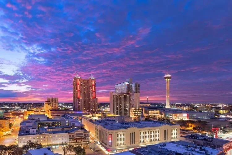 Downtown San Antonio skyline at sunset with colorful purple and pink sky, featuring high-rise buildings and the Tower Life Building.