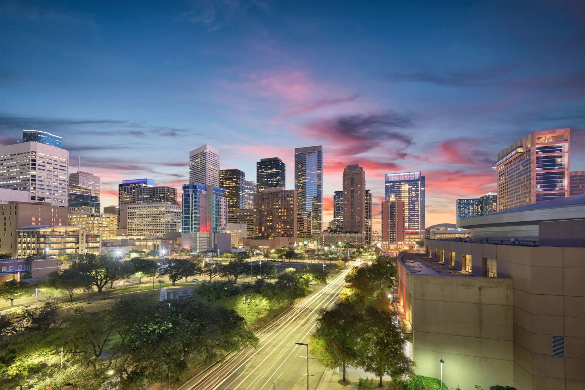 City skyline at dusk with tall modern buildings, colorful sunset sky, and light trails from moving vehicles on the street.