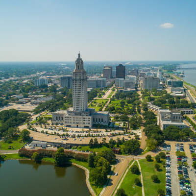 Aerial view of a cityscape featuring a tall historic building with a clock tower, surrounded by modern office buildings, parks, and a lake on a sunny day.