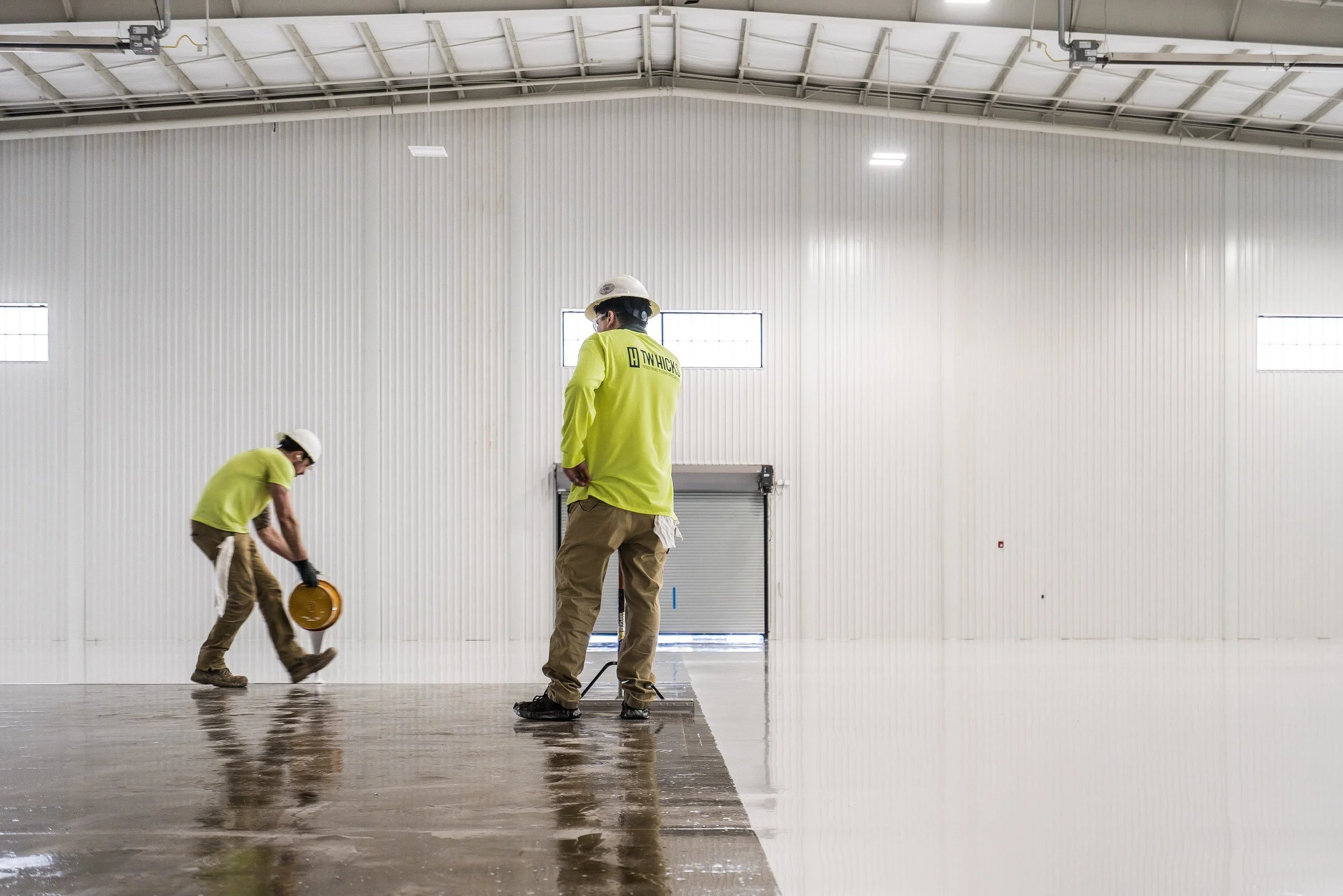 Construction workers in high-visibility clothing and hard hats working in an indoor space with a high ceiling, white walls, and a partly reflective floor.