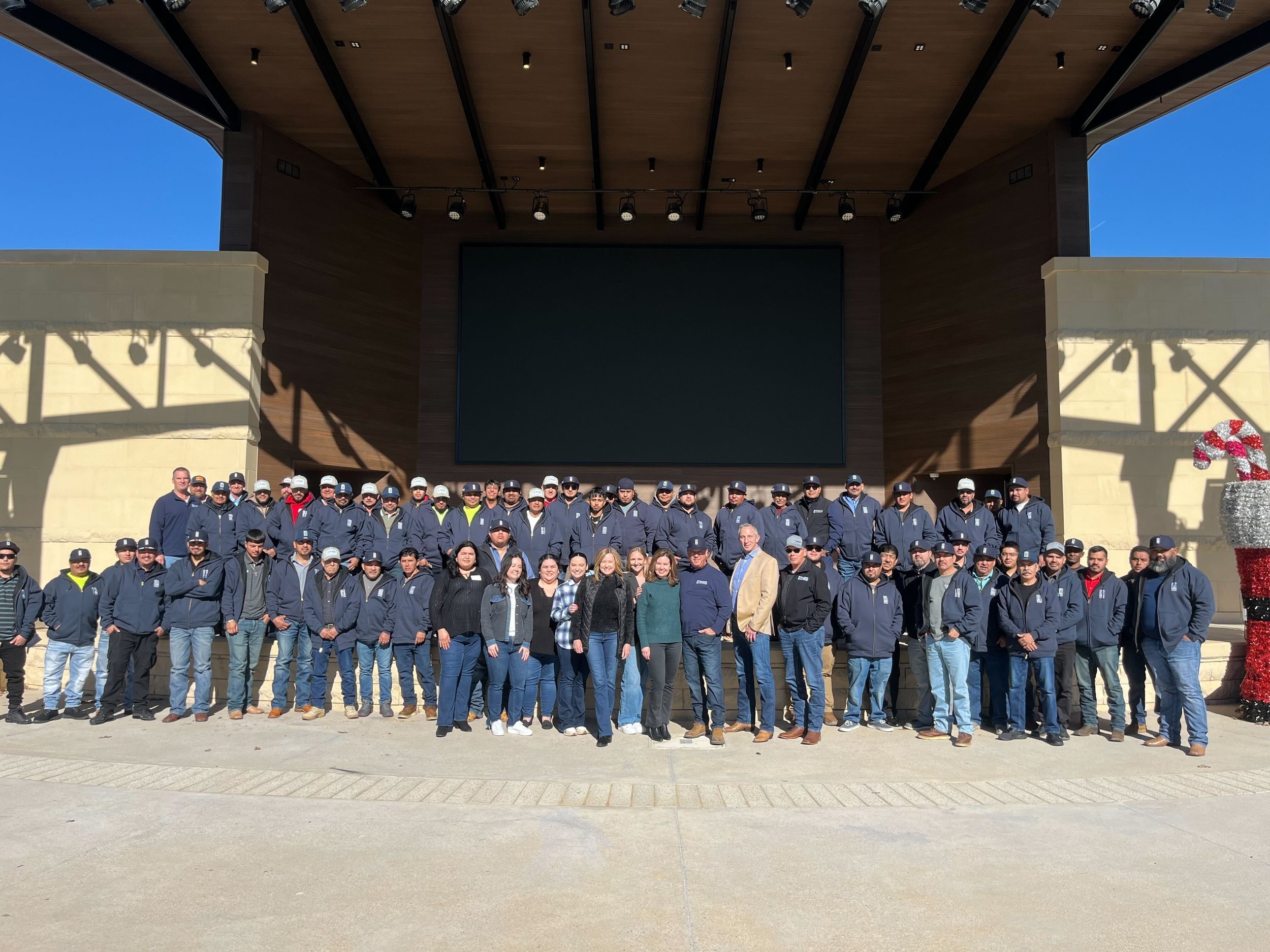 A large group of people standing outdoors in front of a stage with a large black screen and wood-paneled ceiling. The group includes men and women wearing dark jackets and baseball caps, with some smiling and posing for the photo. The setting appears to be a formal or ceremonial gathering.