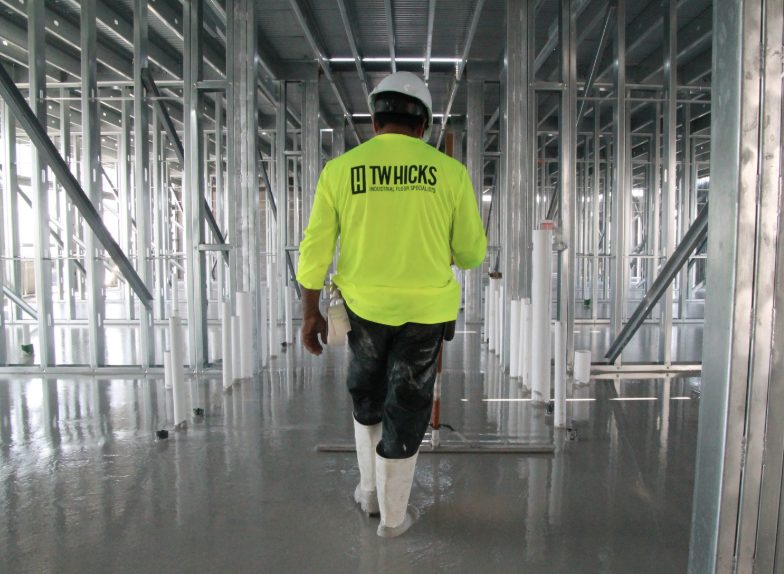 Construction worker walking through a building under construction with metal framing, wearing a yellow shirt, white helmet, and white boots.