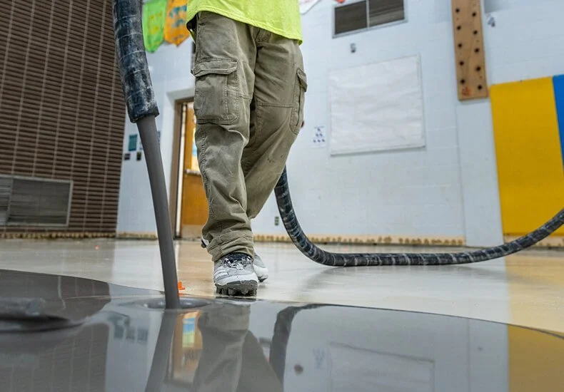 A person in khaki cargo pants and a yellow shirt working with a large industrial vacuum hose on a polished concrete floor.