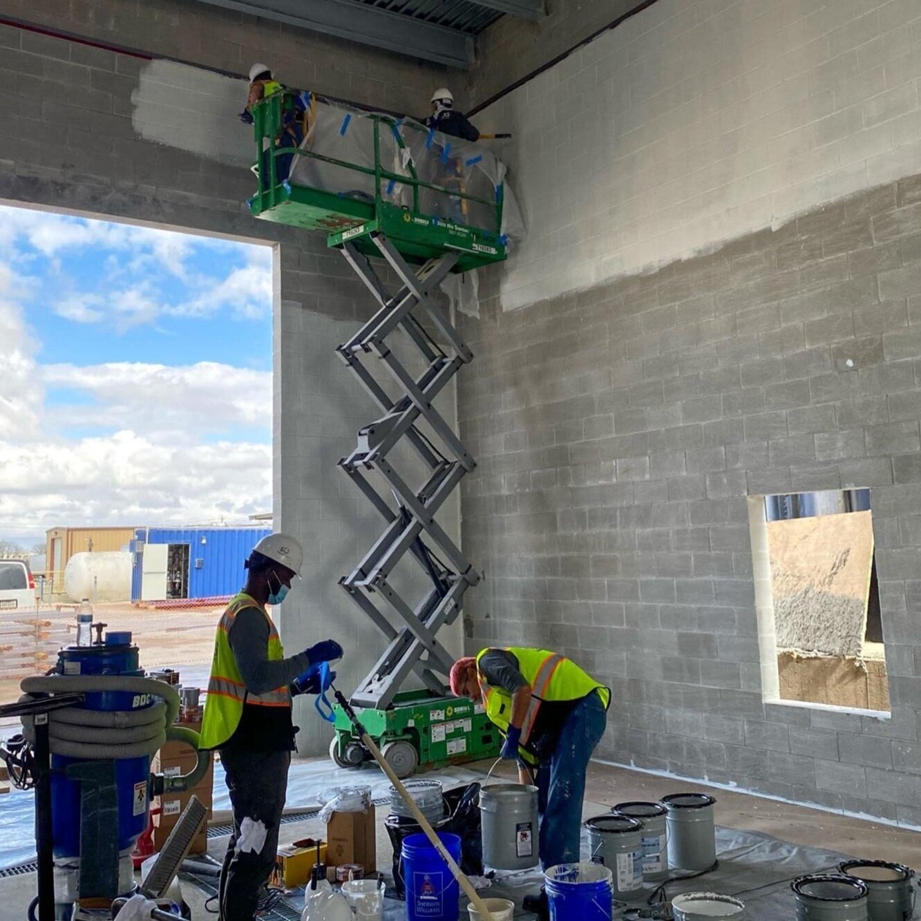 Construction workers in safety gear working on interior wall of building, with a scissor lift elevated to the ceiling. Construction materials and tools scattered on the floor are visible.