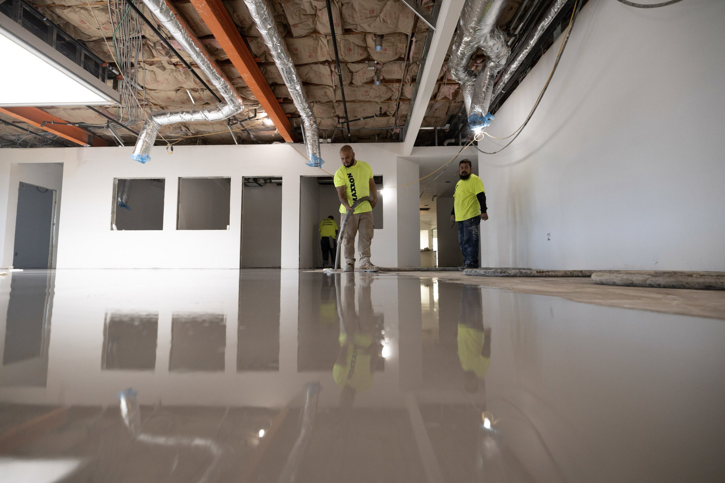 Construction workers installing flooring in a room with drywall and exposed ceiling pipes.