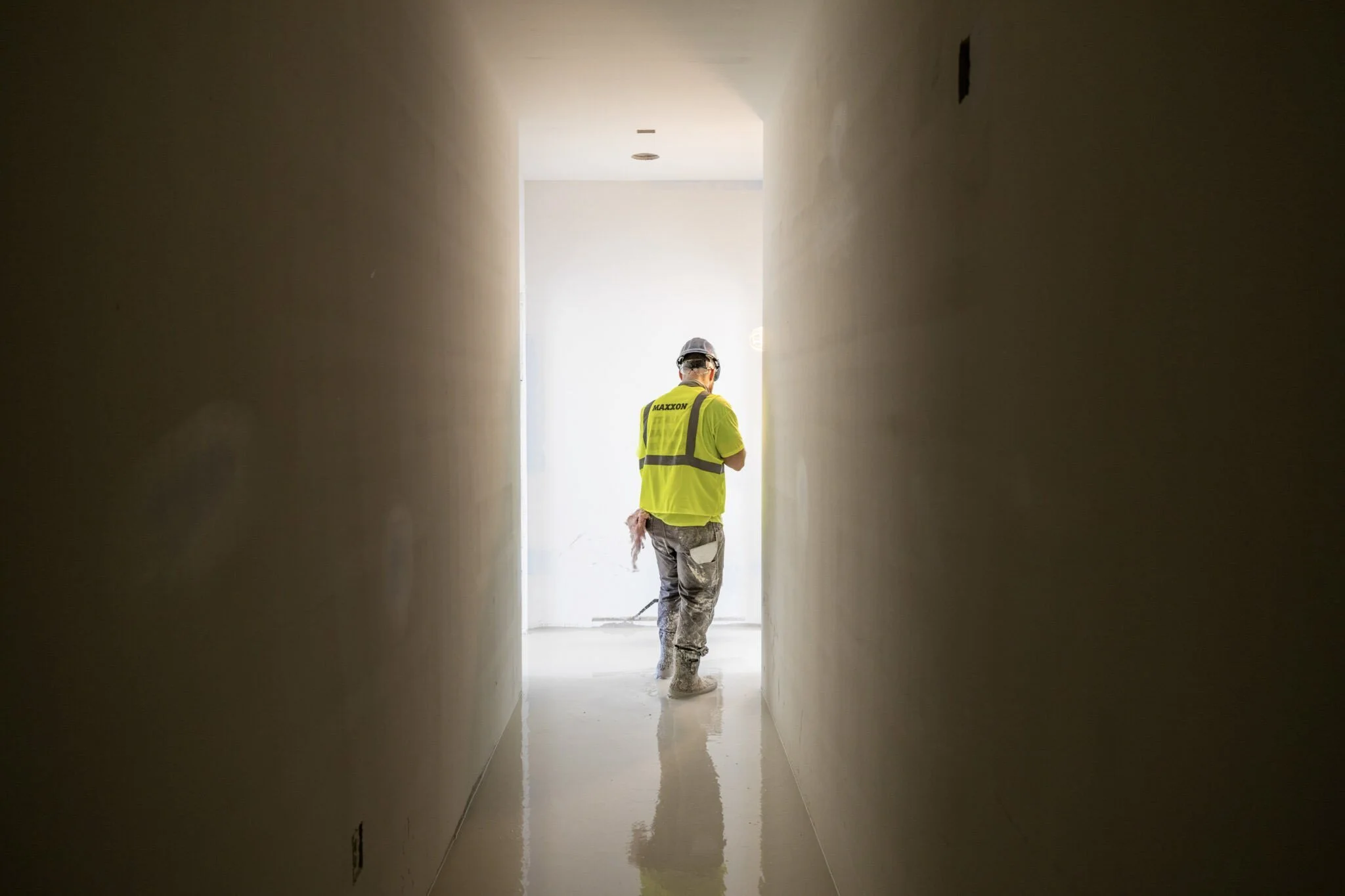 A construction worker wearing a neon yellow safety vest and a hard hat walking through a narrow hallway under construction, illuminated by daylight from outside.