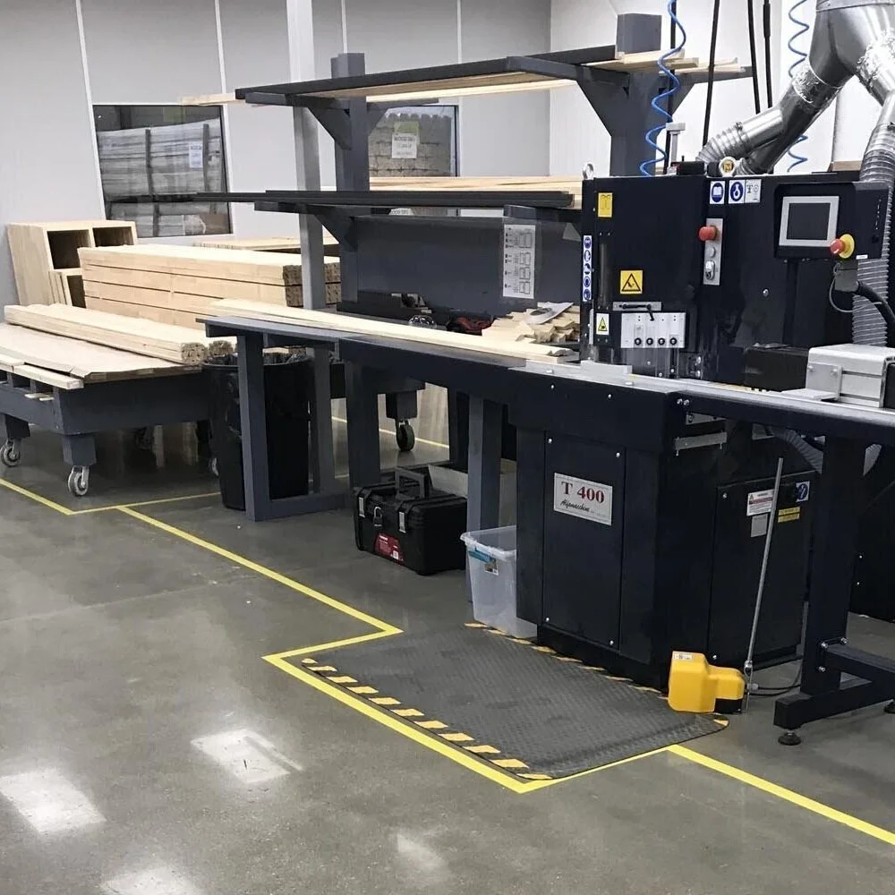 A woodworking workshop with a black industrial woodworking machine, wooden planks, and a workbench. The area is marked with yellow tape on the floor for safety.