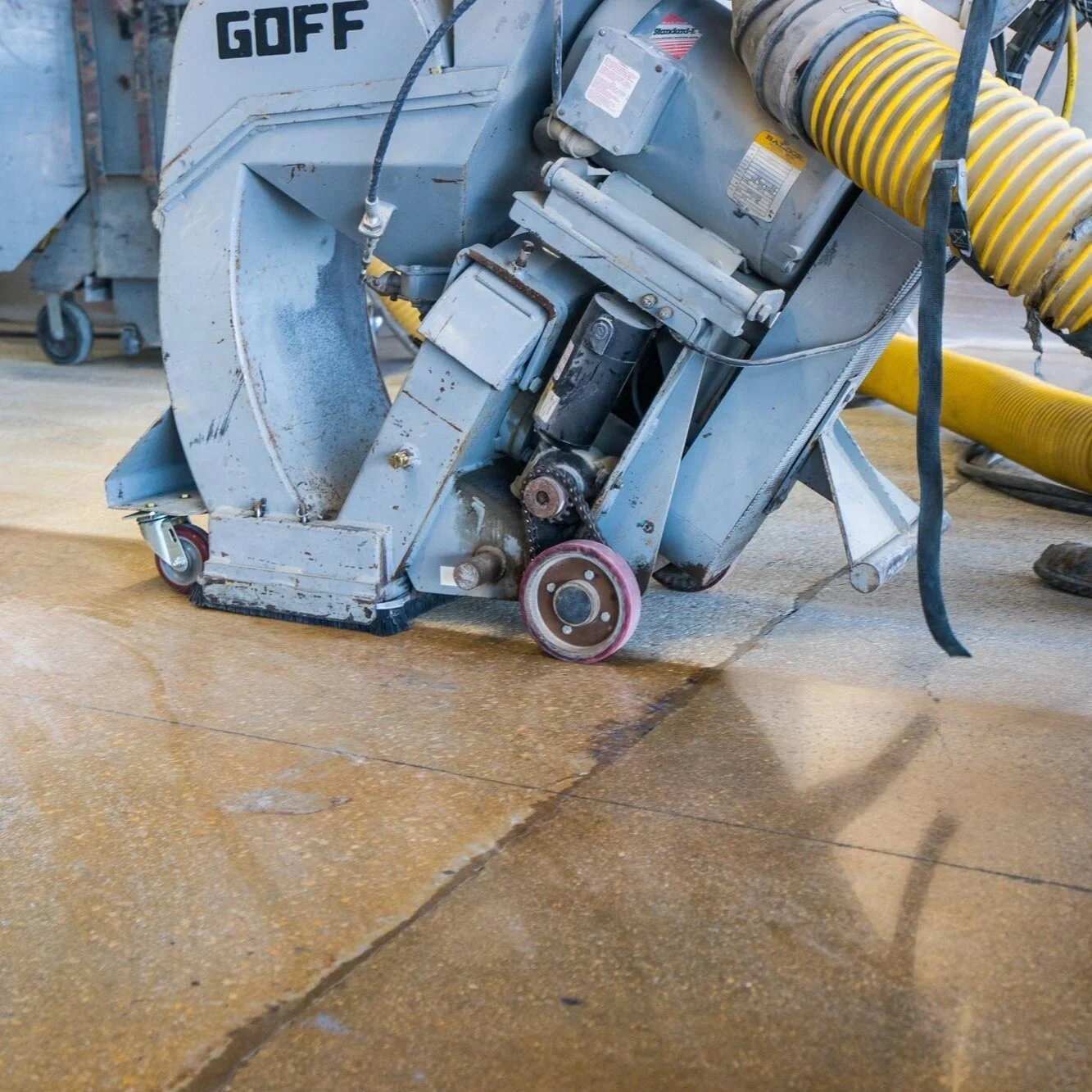 Industrial floor grinding machine on a concrete floor with a yellow vacuum hose attached.