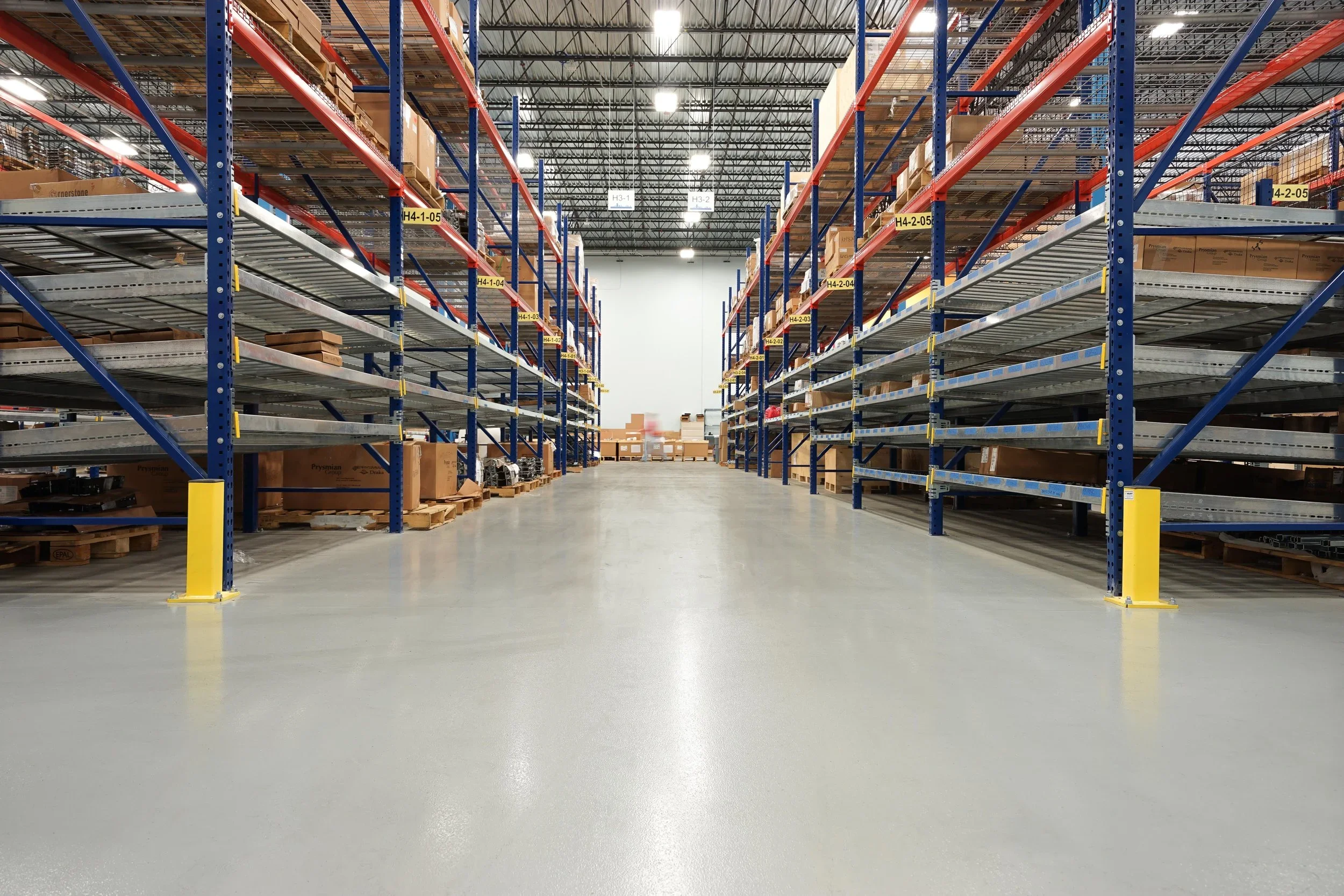 Interior of a warehouse with empty metal shelving units, some with cardboard boxes, and a clear aisle down the middle with a smooth concrete floor.