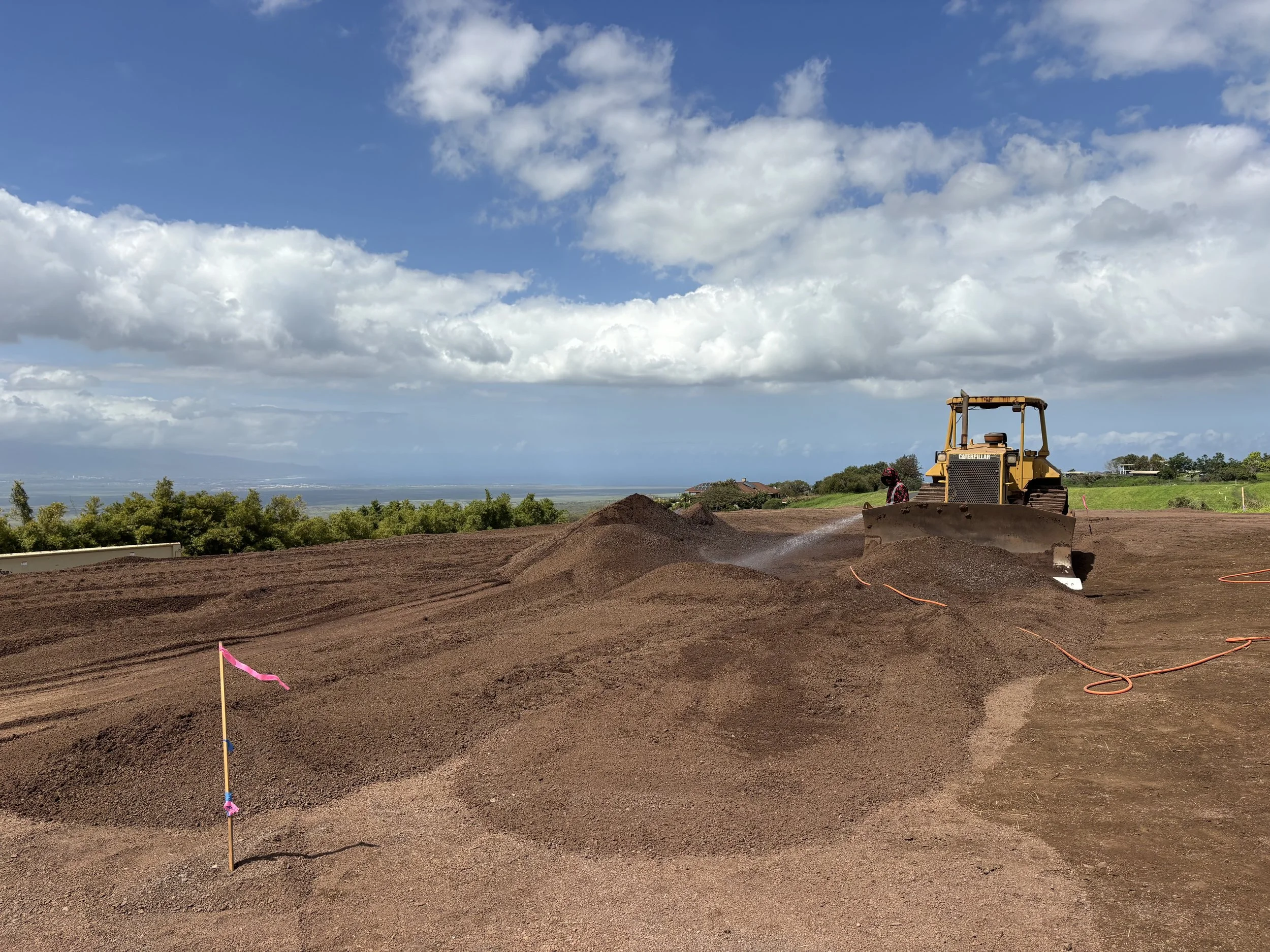 A construction site with a bulldozer on the right side, pink flags marking the area, and a dirt pathway. In the background, there are trees and a partly cloudy sky.