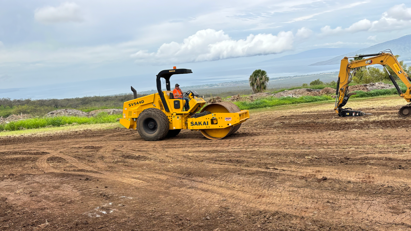 A construction site with heavy machinery, including a yellow steamroller and an excavator, on a dirt road with a scenic landscape and lake in the background.