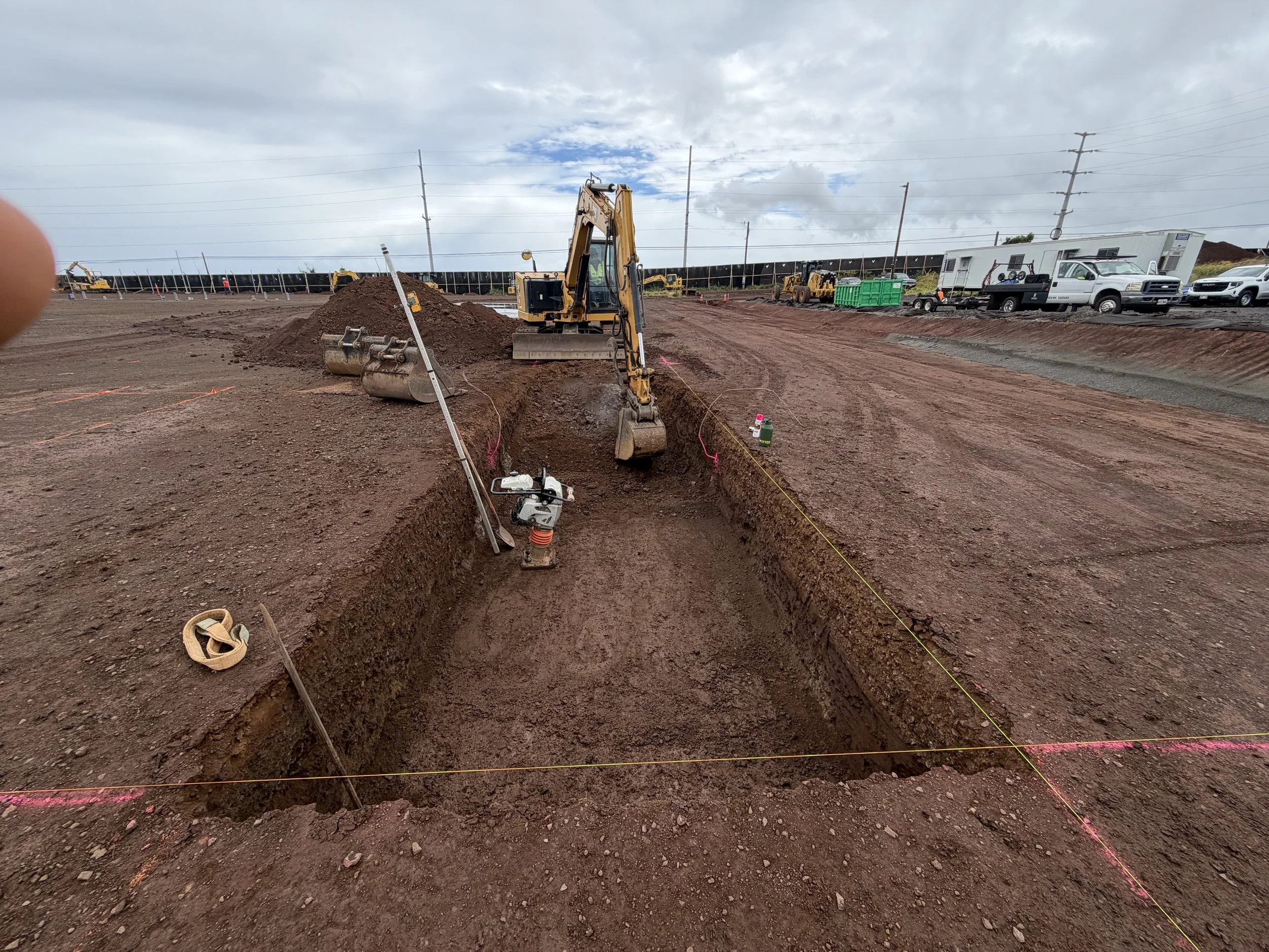 Construction site with dirt excavation, yellow mini excavator, construction vehicles, and utility vehicles under cloudy sky.