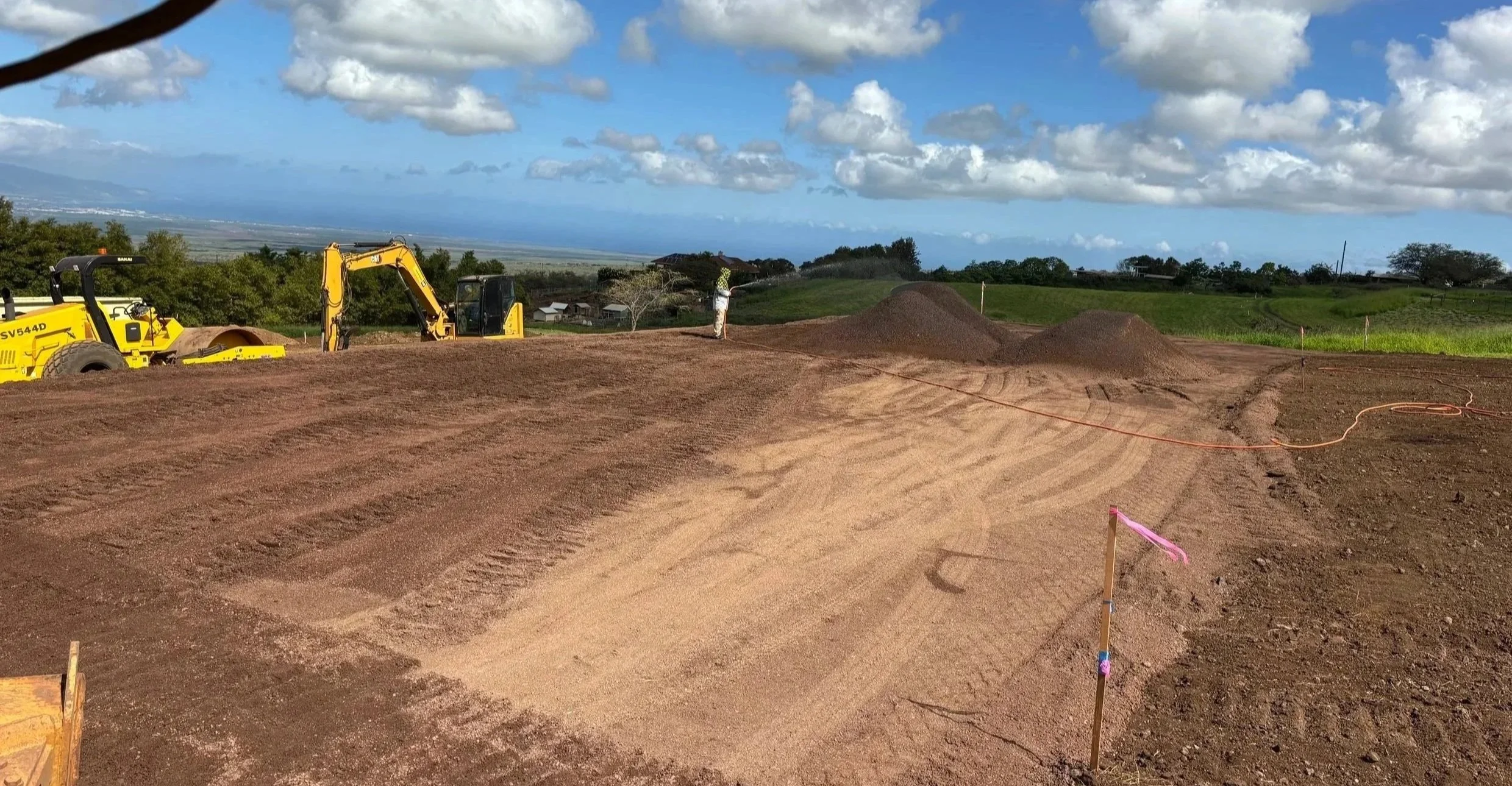 Construction site with yellow machinery and a person working on the dirt, with green fields and a partly cloudy sky in the background.