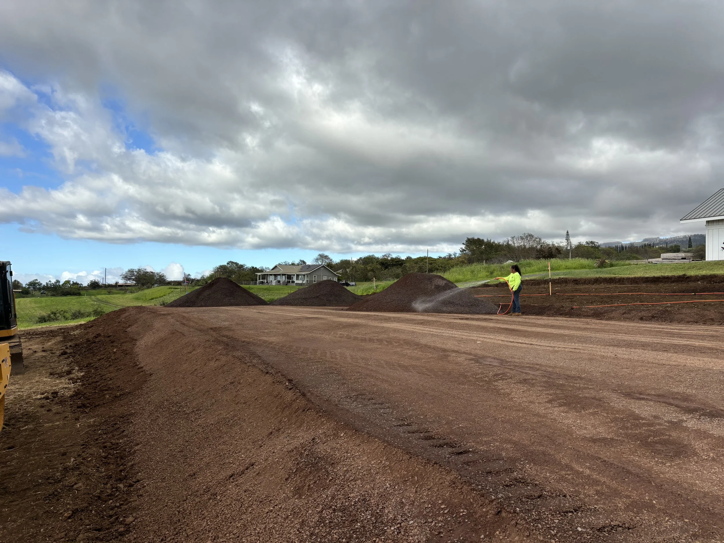 A person watering newly laid soil on a construction site in a rural area with houses and trees in the background, under a cloudy sky.