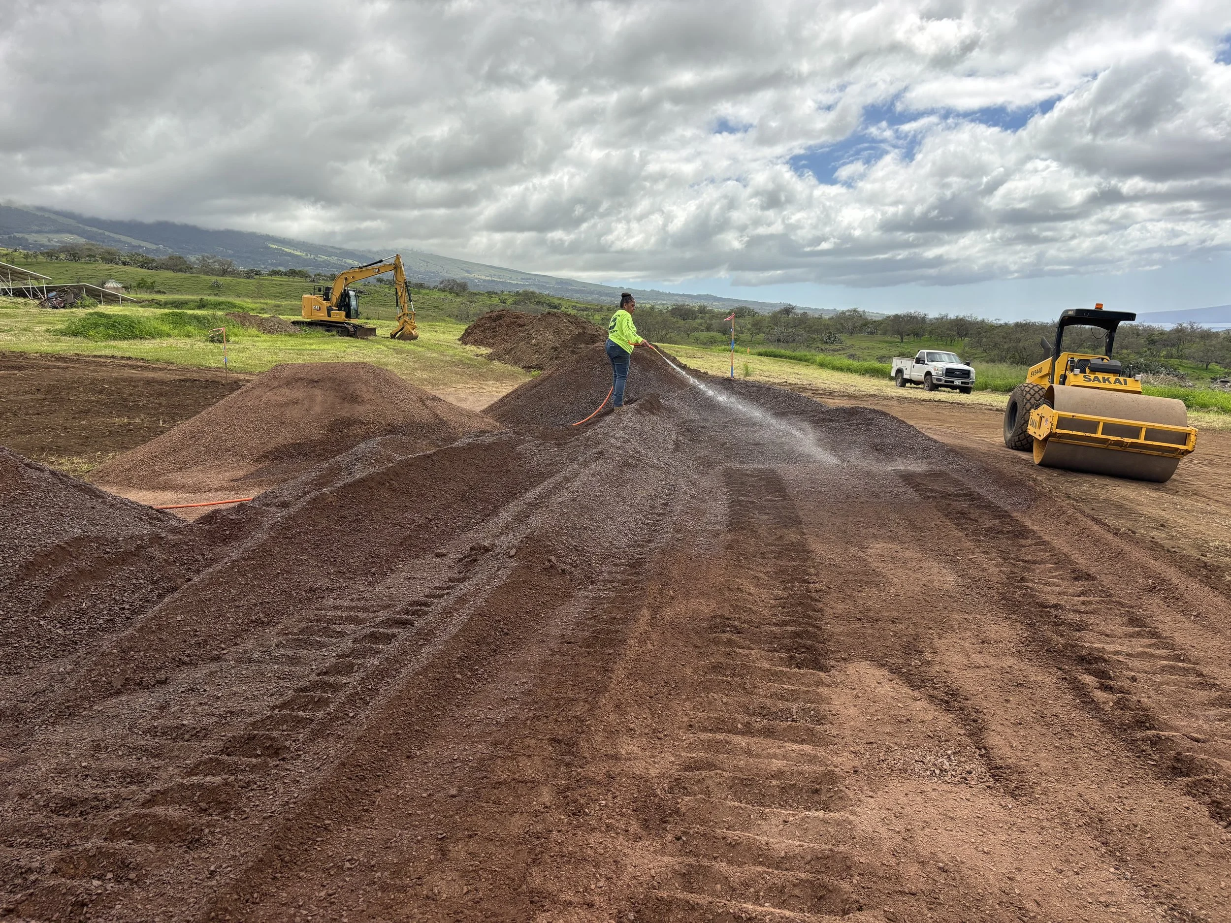 A person watering large piles of reddish soil or mulch with a garden hose on a construction or landscaping site. Construction equipment and a vehicle are visible in the background under cloudy skies.