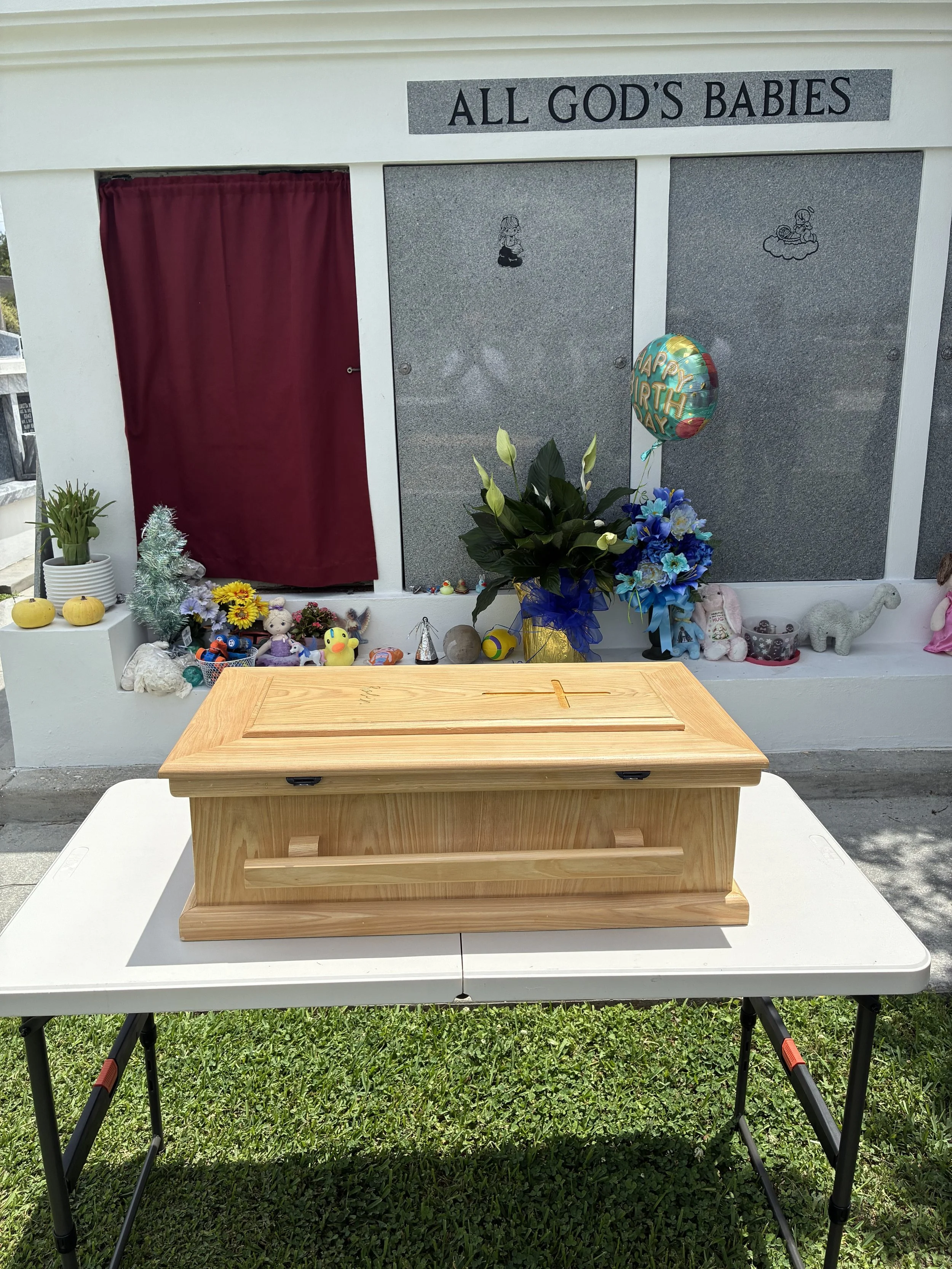 A grave with a wooden box and a cross on top, surrounded by toys, flowers, and decorations, with a memorial display behind it that reads 'ALL GOD'S BABIES' and features illustrations of children and a baby in a cradle. There are balloons and plants n