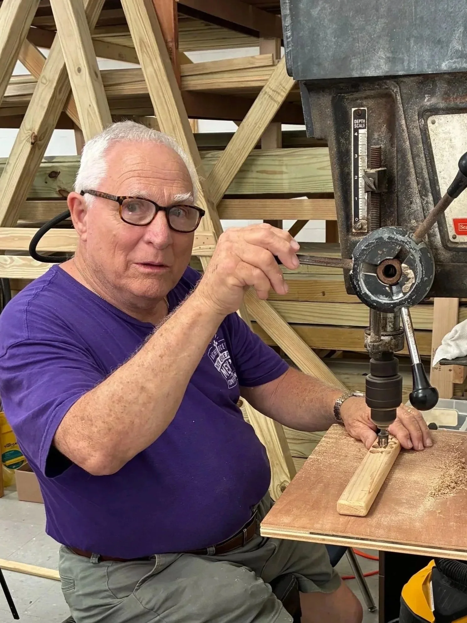 An elderly man with gray hair and glasses working with a drill on a piece of wood in a woodworking workshop. There are wooden structures in the background.