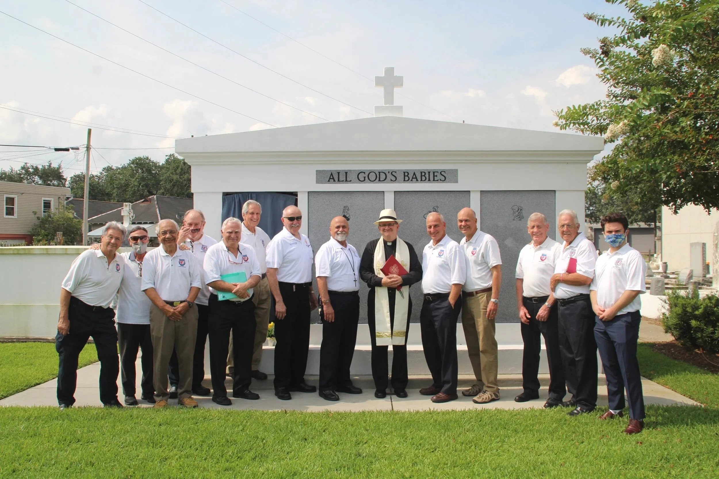 Group of men and women standing in front of a white monument with the inscription 'All God's Babies', with a priest or minister in the middle wearing religious garments and holding a book, outdoors on a sunny day.