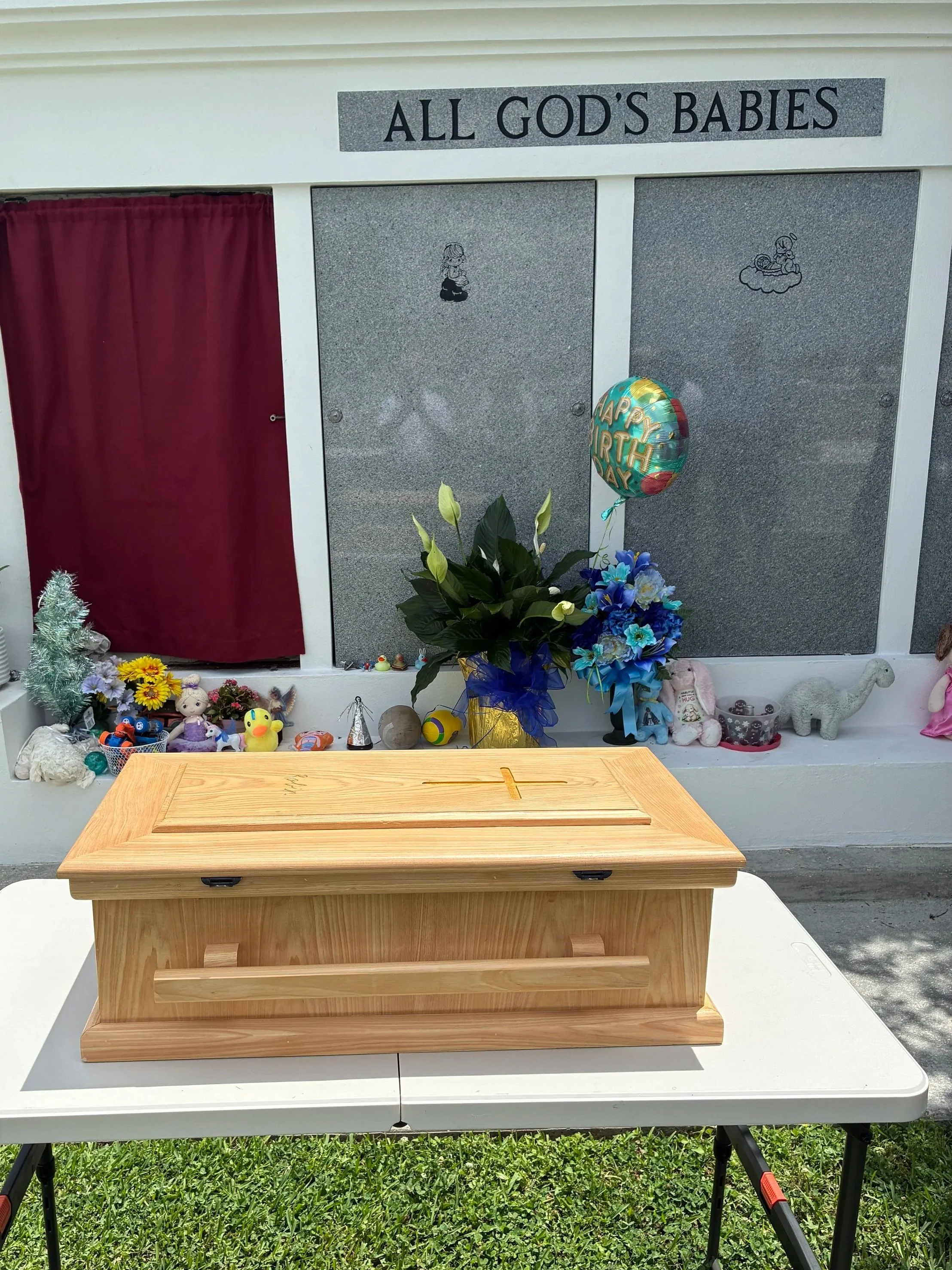A wooden urn on a white table, with a red curtain behind it, surrounded by various stuffed animals, toys, and decorative items. A balloon with a colorful design and the words "Happy Birthday" is affixed to a plant with white flowers next to the urn.