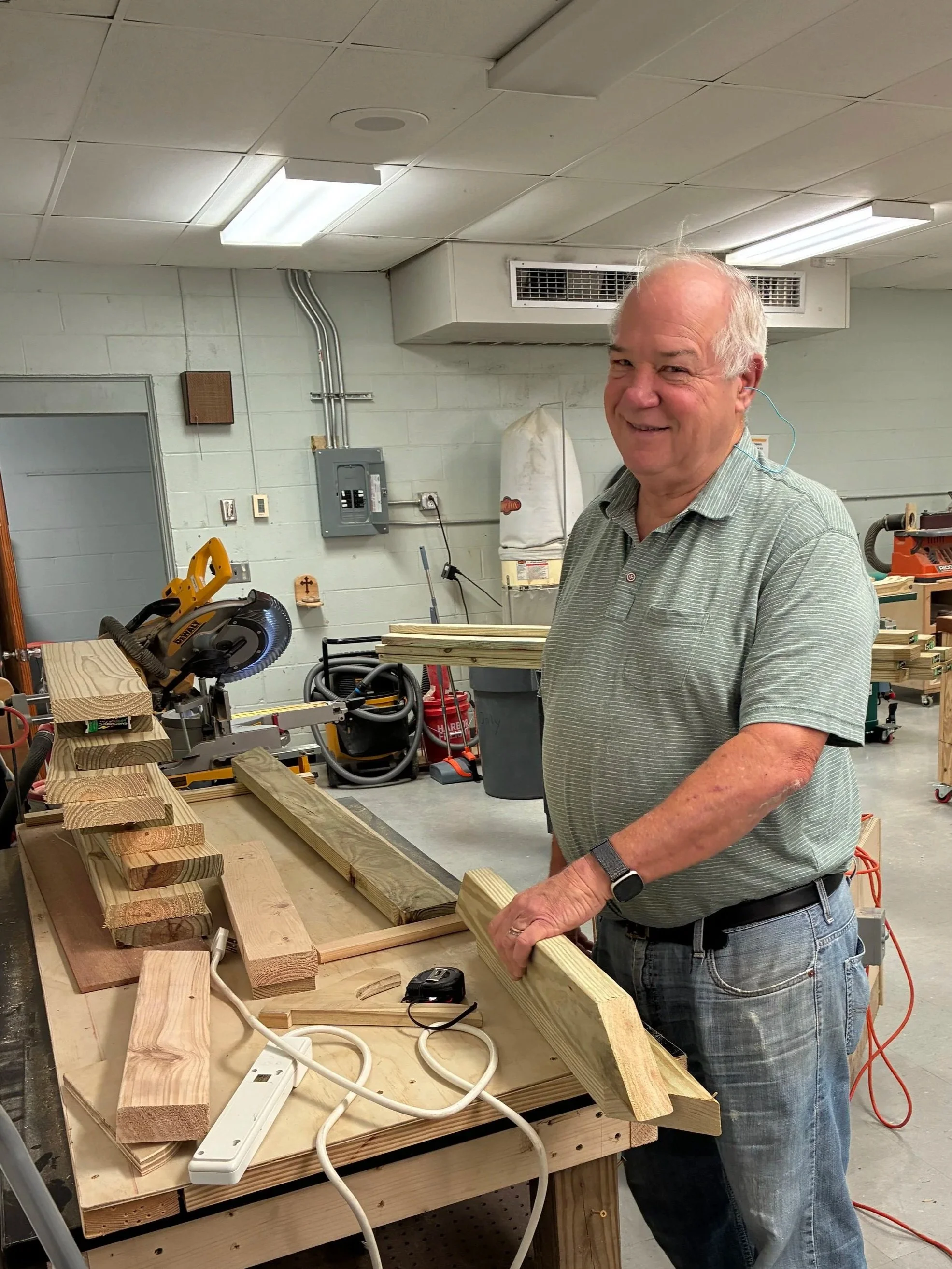 A man in a green striped shirt and jeans working in a woodworking workshop, holding a piece of wood, with woodworking tools and pieces of wood on a workbench.