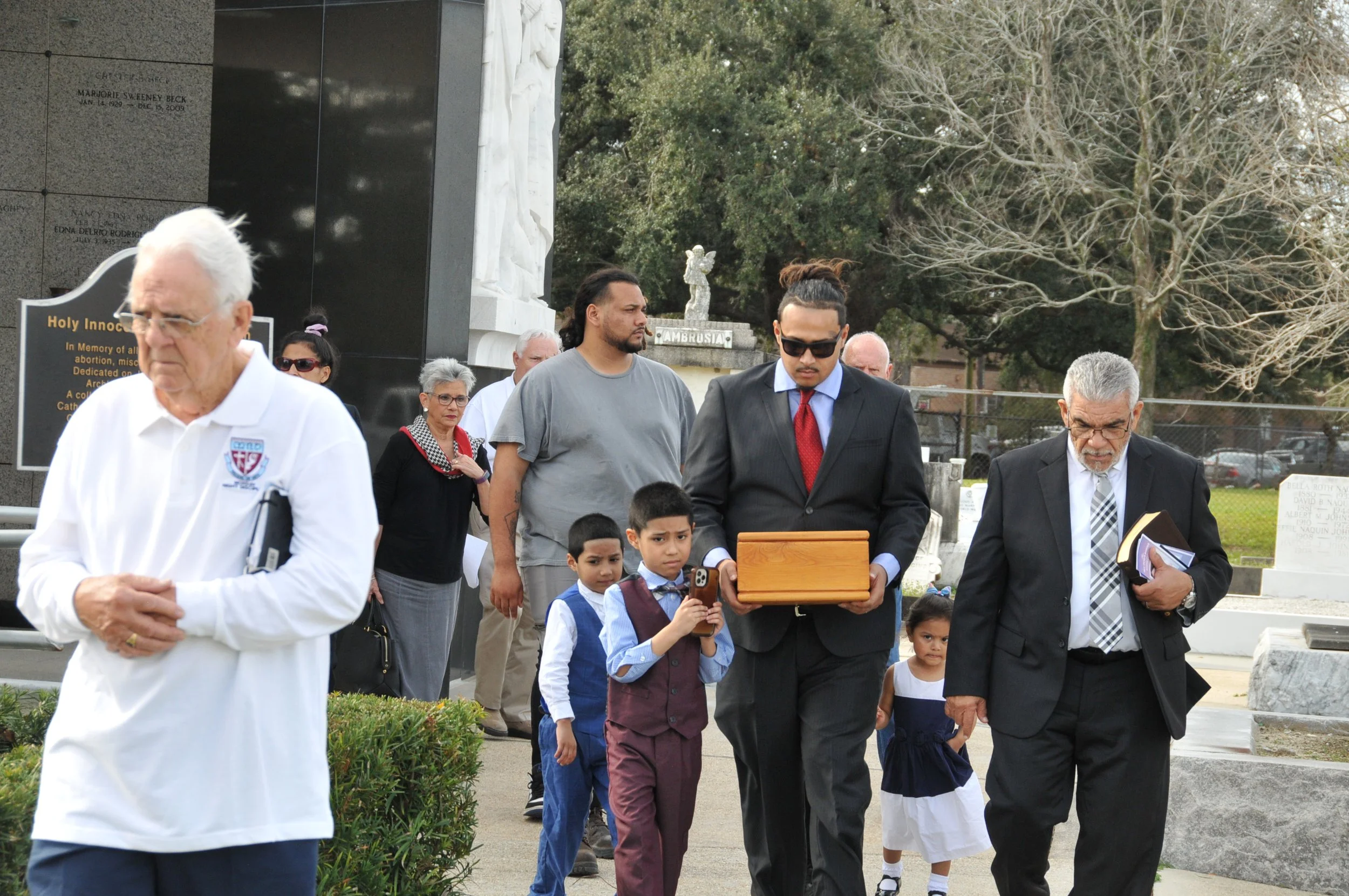 A diverse group of people, including children and adults, gathered in a cemetery, paying respects at a memorial site with statues and engraved plaques, outdoors with trees in the background.