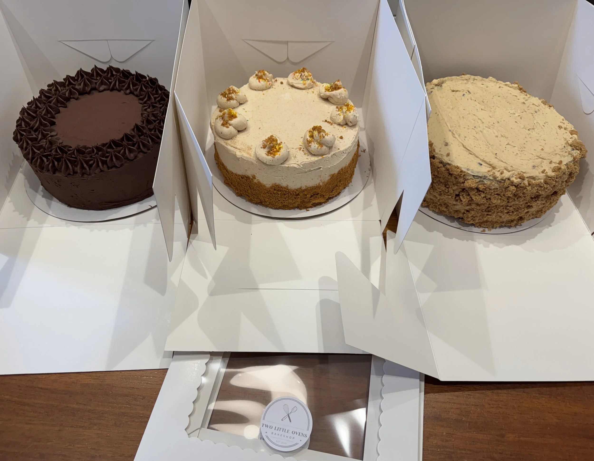 Three different cakes in white bakery boxes. The first cake is chocolate with piped frosting around the top edge. The second cake is white with dollops of cream and yellow-orange sprinkles on top. The third cake is light brown with a crumbly texture.