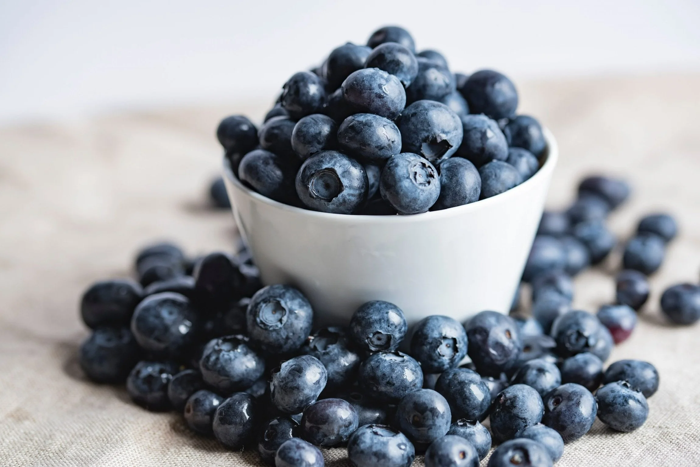 Fresh blueberries in a white bowl with some spilled on a beige fabric surface.