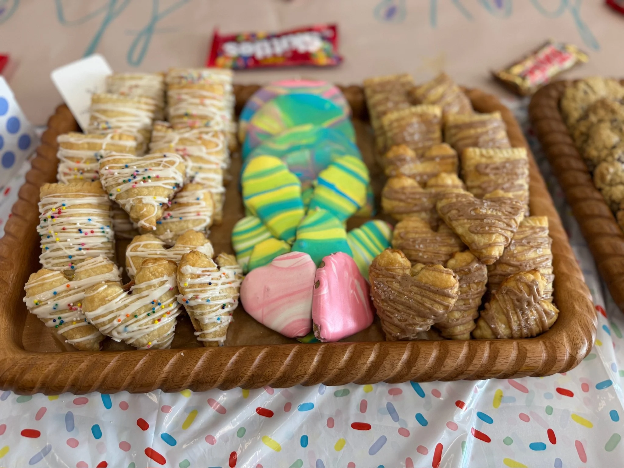 Assorted cookies and treats, including pinwheel cookies with colorful icing, chocolate-chip cookies, and cookies with rainbow sprinkles on a wooden tray.
